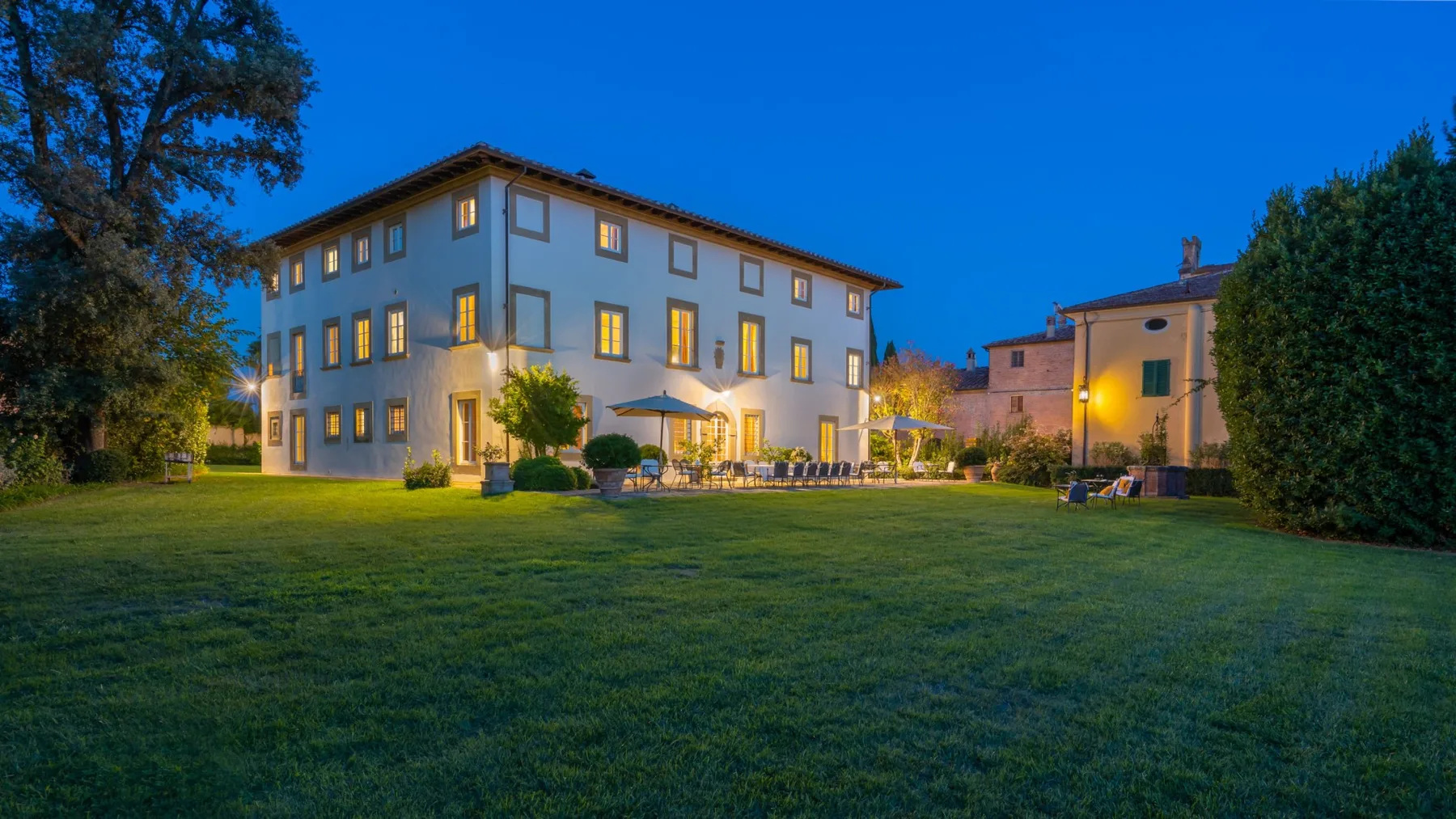 A large building overlooks a grassy lawn at dusk, with outdoor seating beside it.