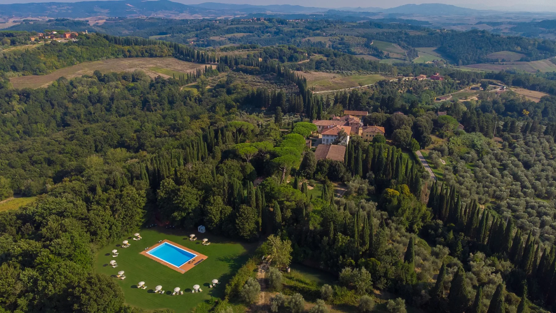 An aerial view shows a swimming pool beside a wooded hillside property.