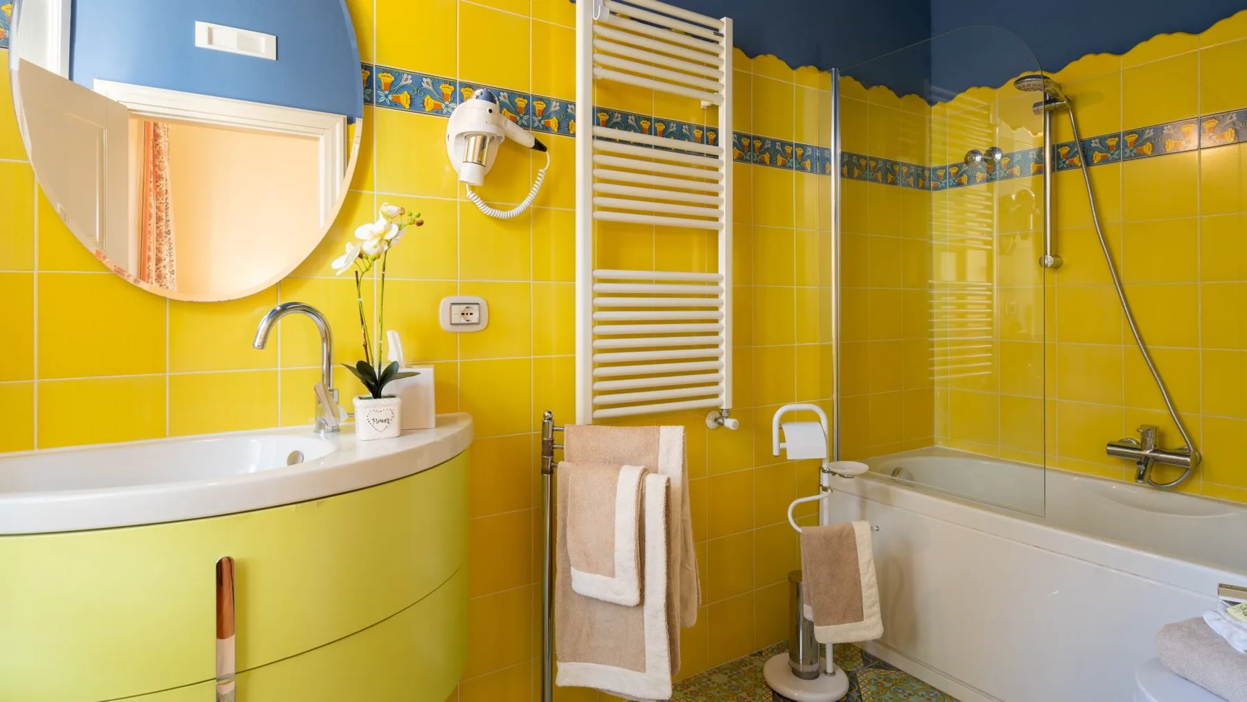 A bright yellow-tiled bathroom with a sink, bathtub, and wall-mounted towel warmer.