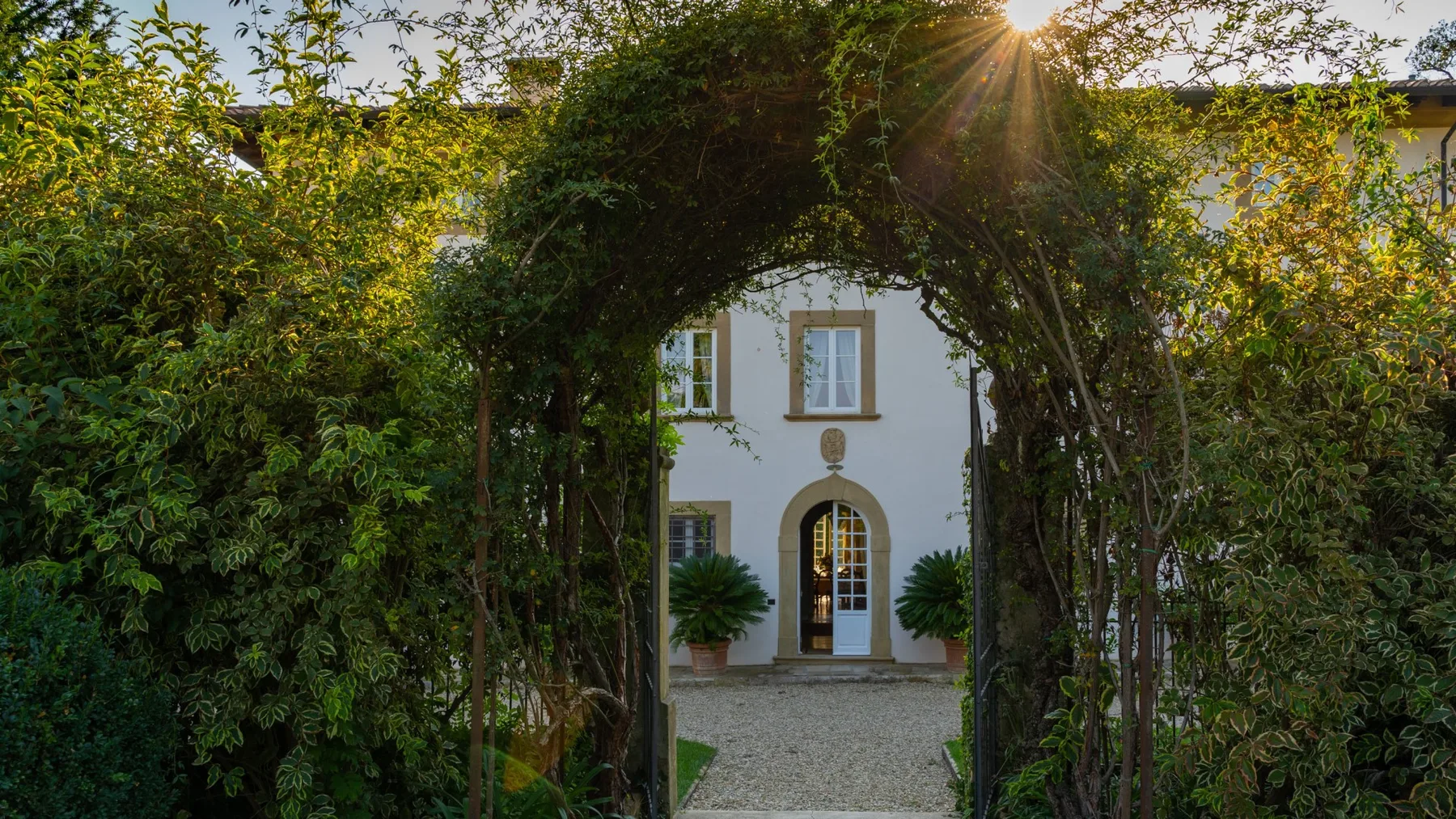 A white house stands beyond a leafy garden arch, with a gravel path leading to its entrance.