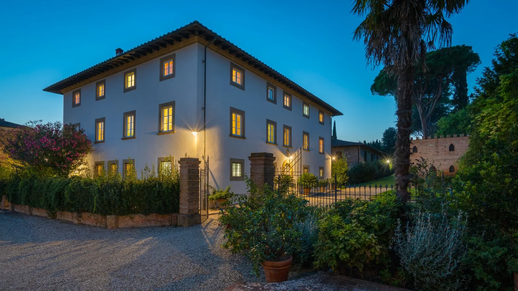 A multi-story building glows with warm window light at dusk, behind a garden and gate.