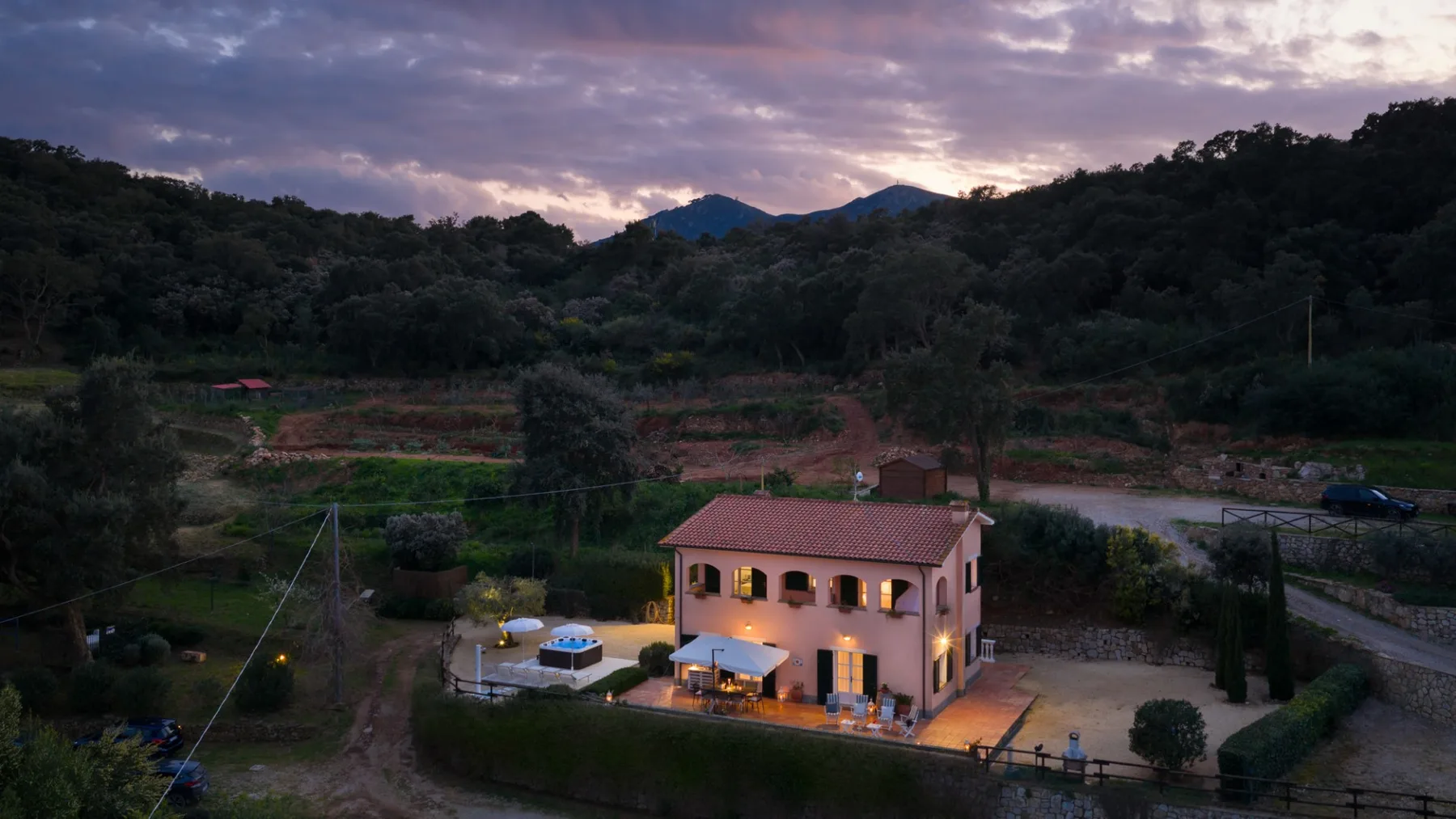A lit two-story house stands among wooded hills beneath a cloudy evening sky.