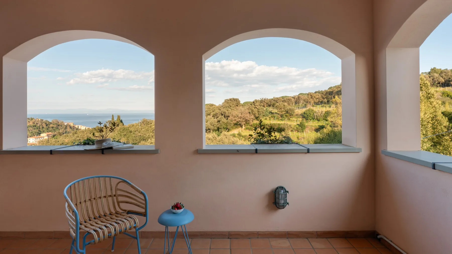A covered terrace has arched openings overlooking the sea and green hills.
