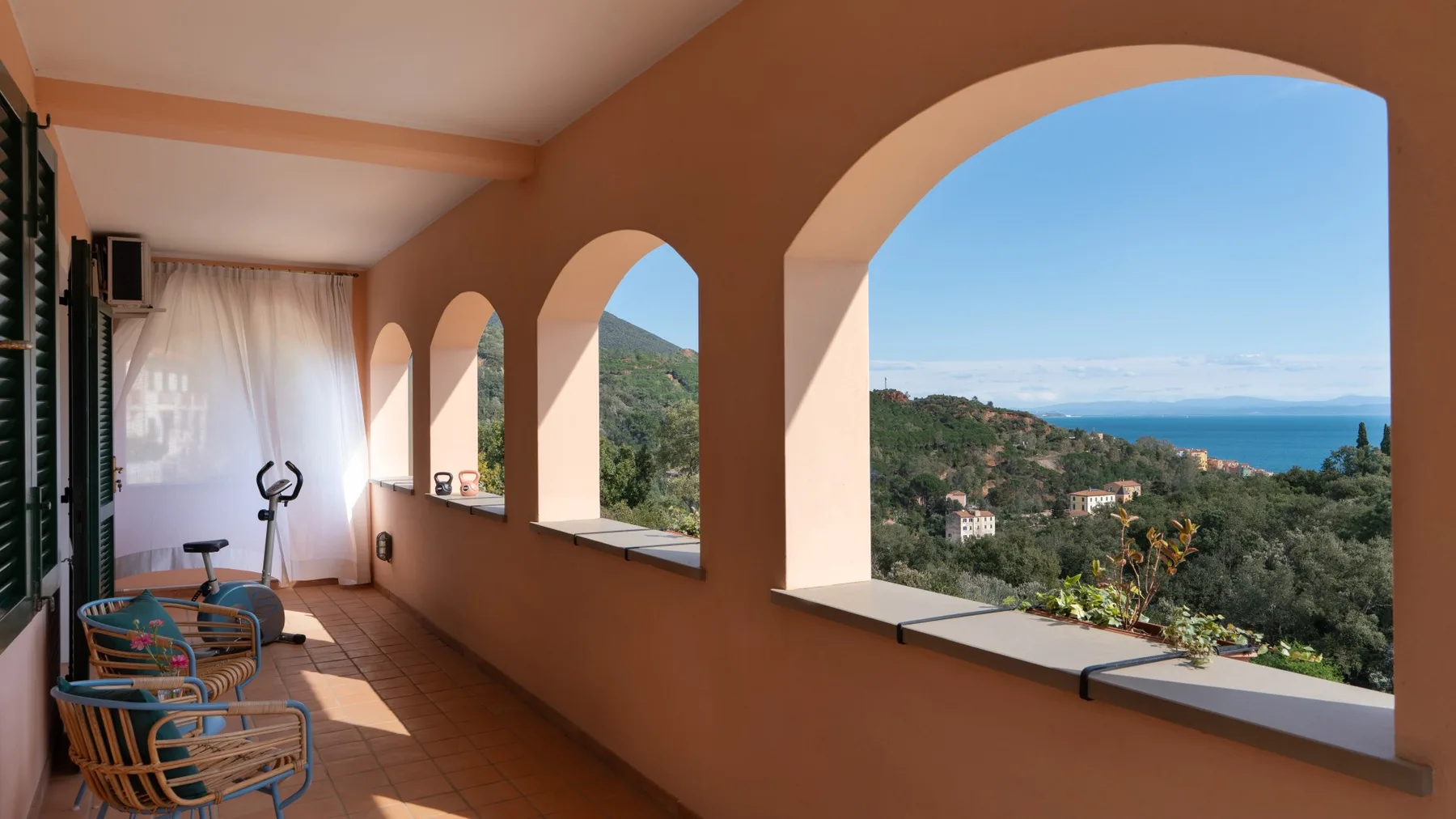 A covered terrace has arched openings overlooking a green hillside and the sea.