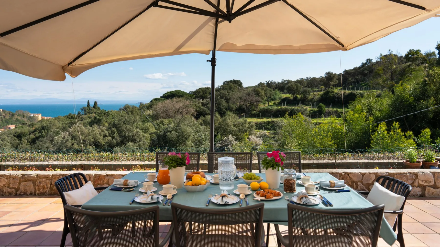 An outdoor dining table sits beneath a large umbrella, overlooking a green hillside and sea.