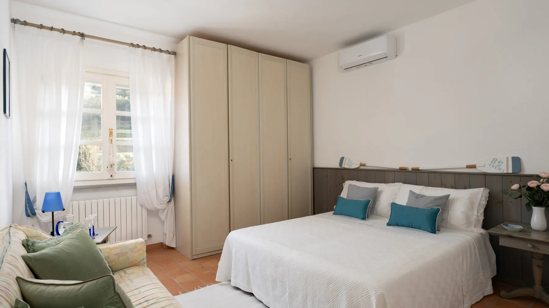 A bedroom with a neatly made bed, wardrobe, and window with sheer curtains.