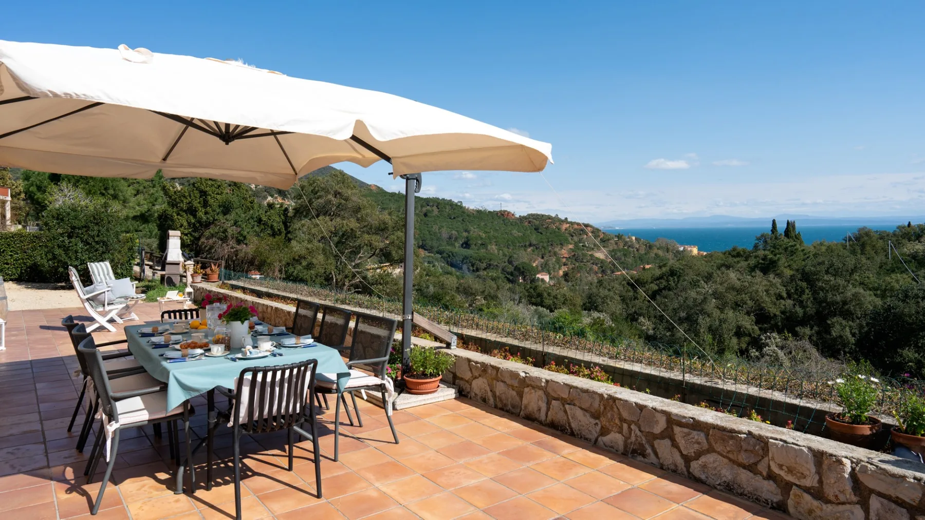 A sunny outdoor dining terrace overlooks green hills and the sea.