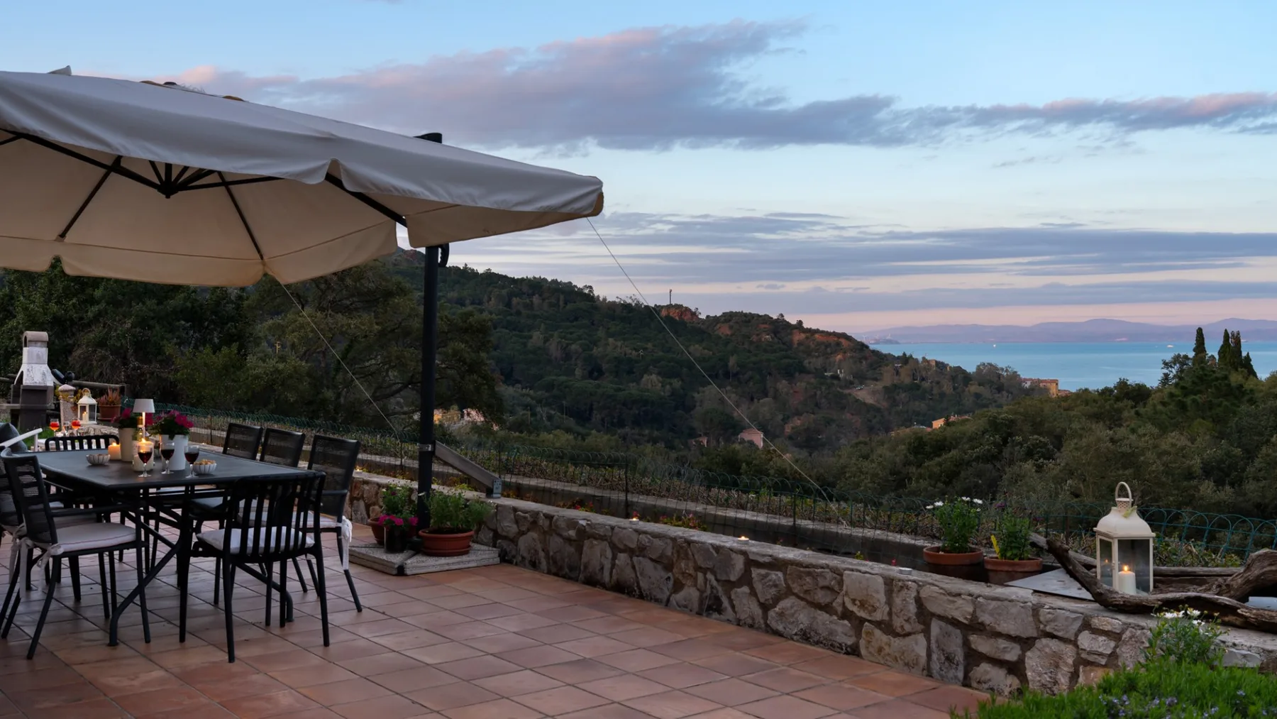An outdoor dining terrace overlooks a wooded hillside beneath a cloudy evening sky.