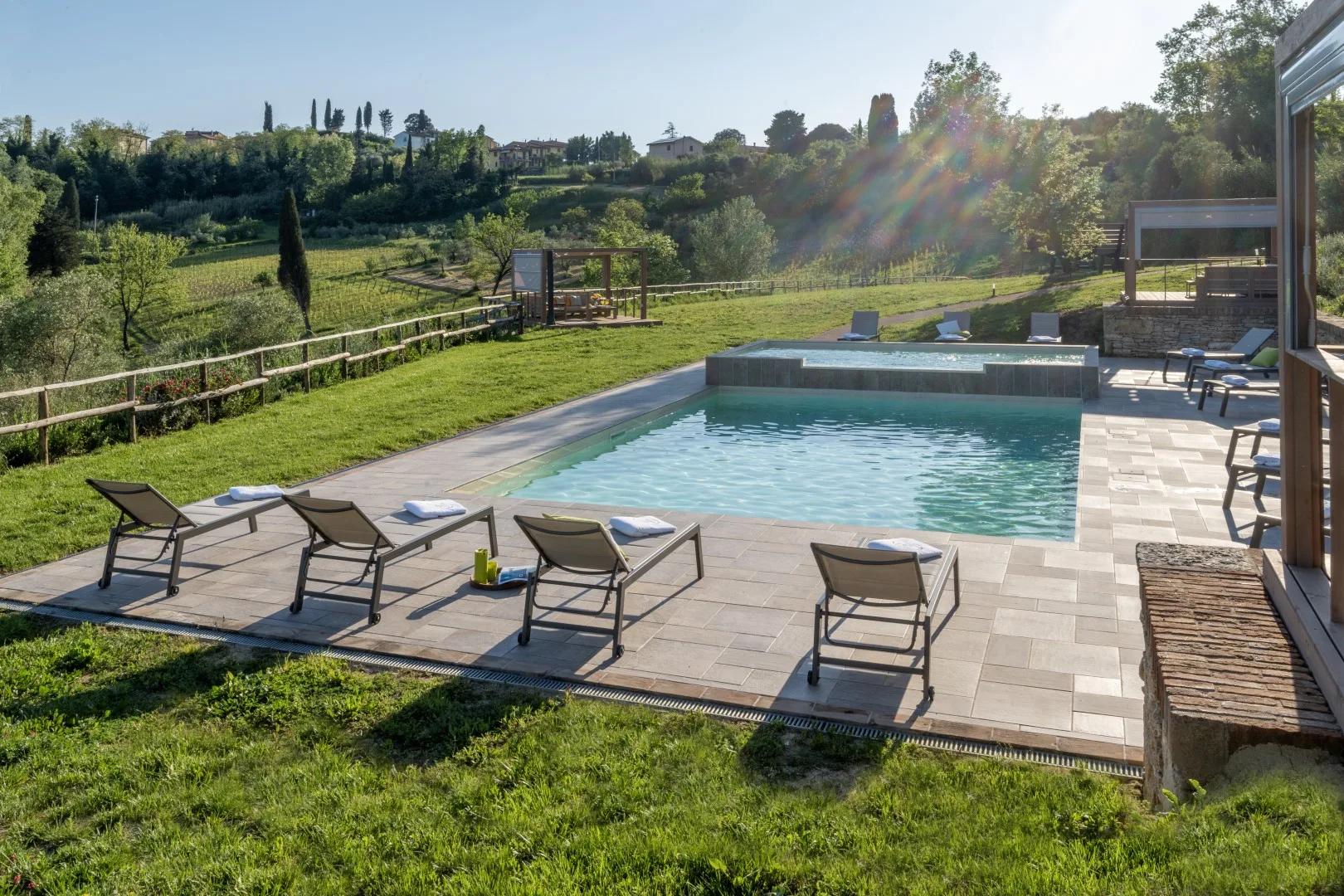 An outdoor swimming pool beside a paved deck with seating and green fields beyond.