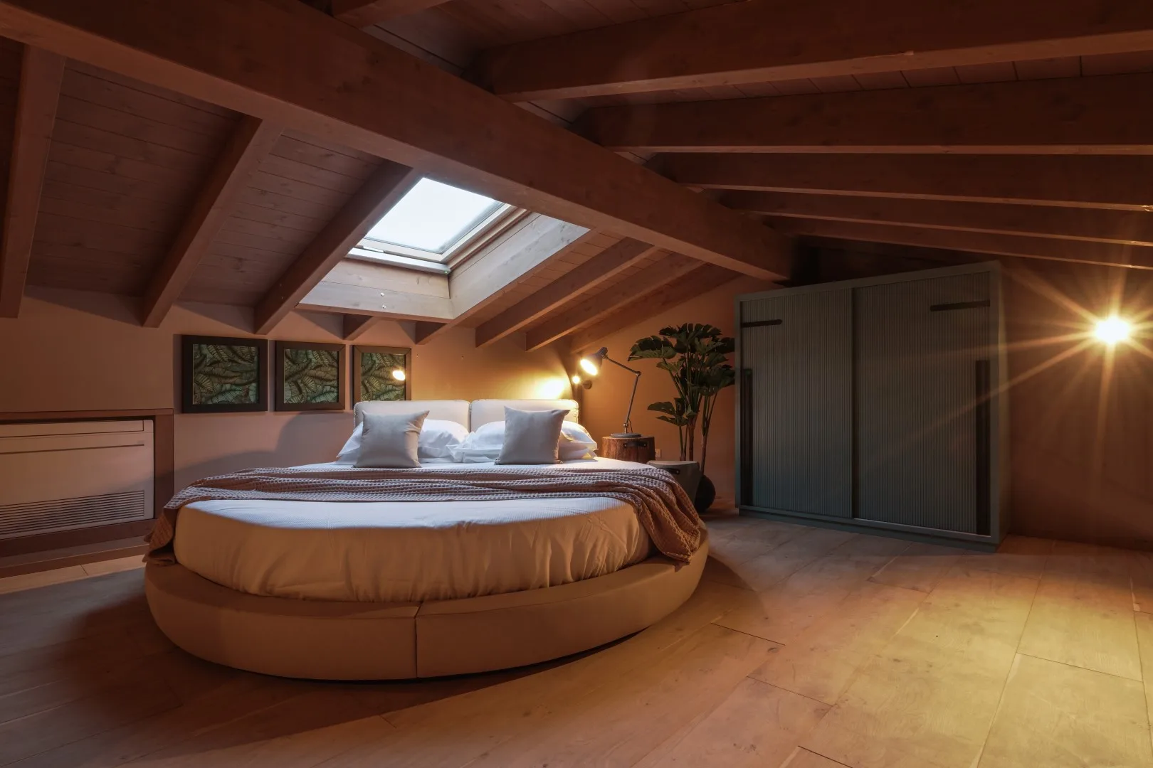 A bedroom with a round bed beneath a sloped wooden ceiling and skylight.