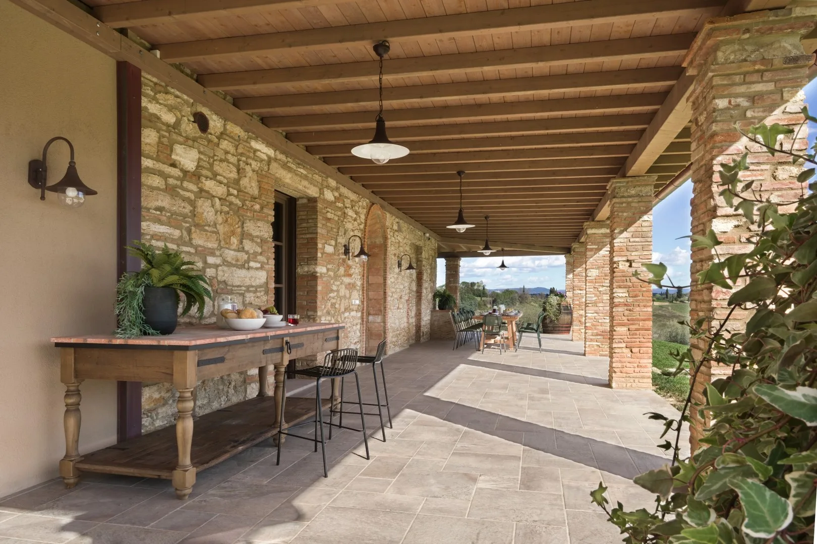 A covered stone veranda with a long table and seating opens onto a garden.