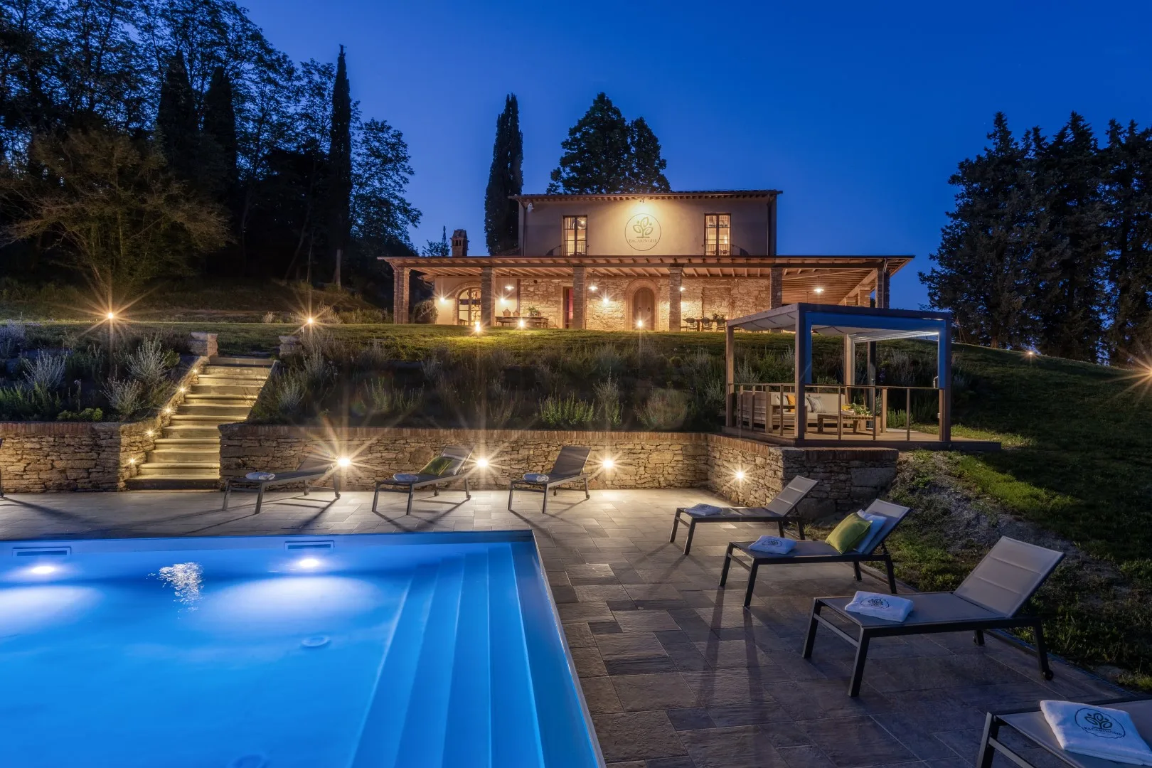 A lit outdoor pool and surrounding patio sit in front of a large house at dusk.