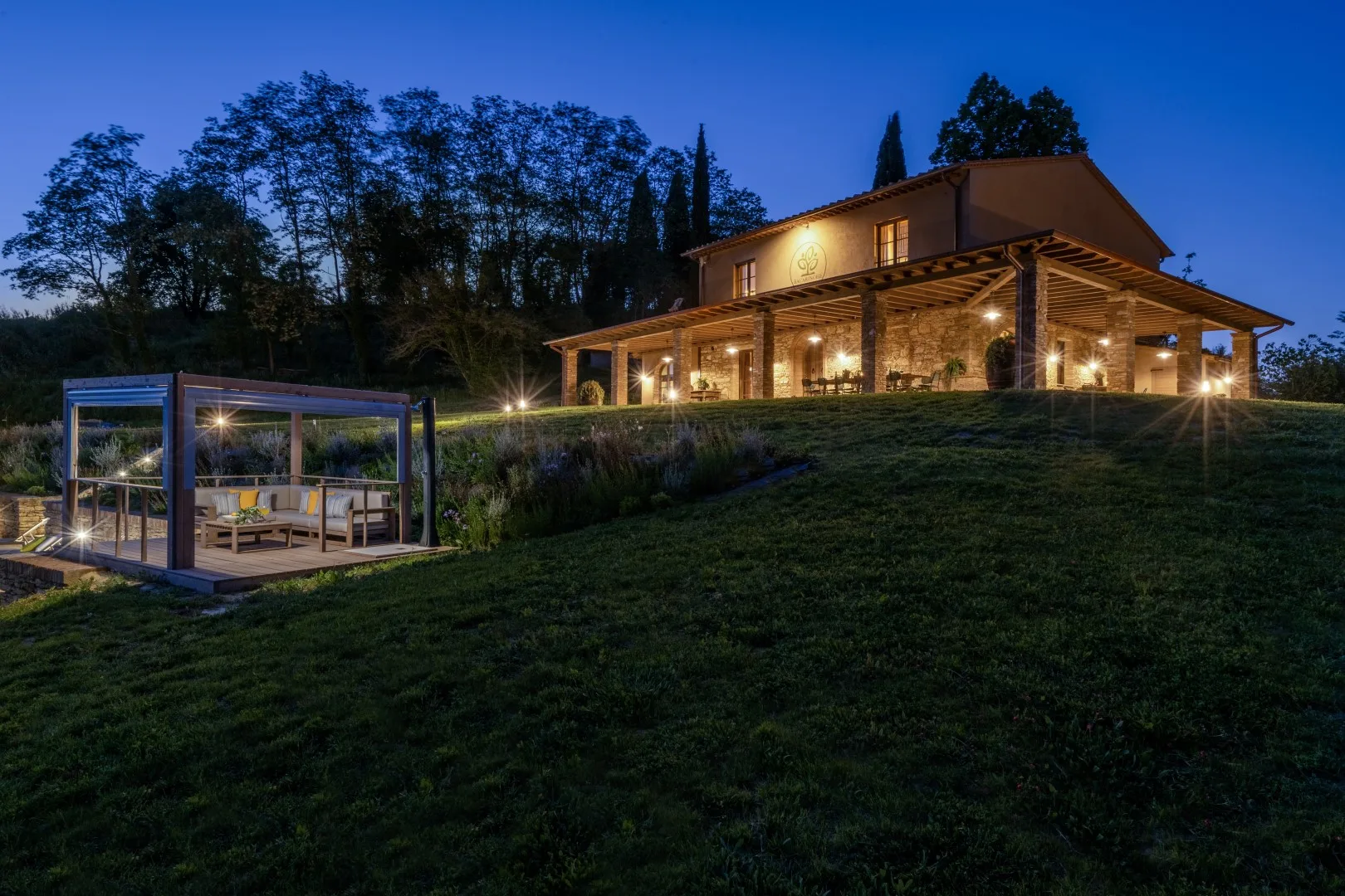 A modern house glows at dusk beside a glass-walled outdoor seating area.