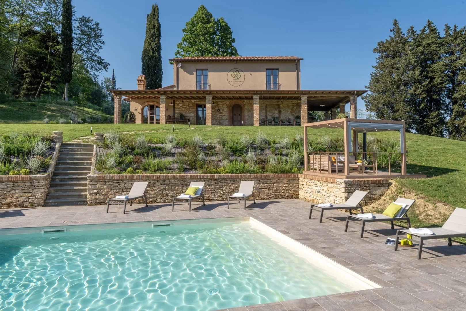 A swimming pool and patio sit in front of a stone terrace and landscaped hillside.