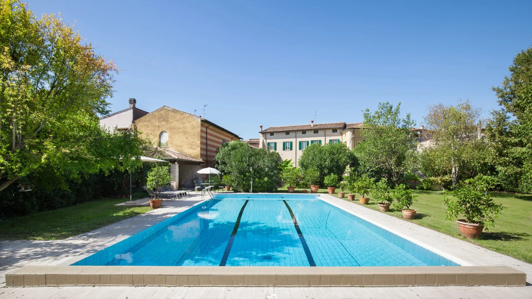 An outdoor swimming pool is surrounded by greenery and neighboring buildings.