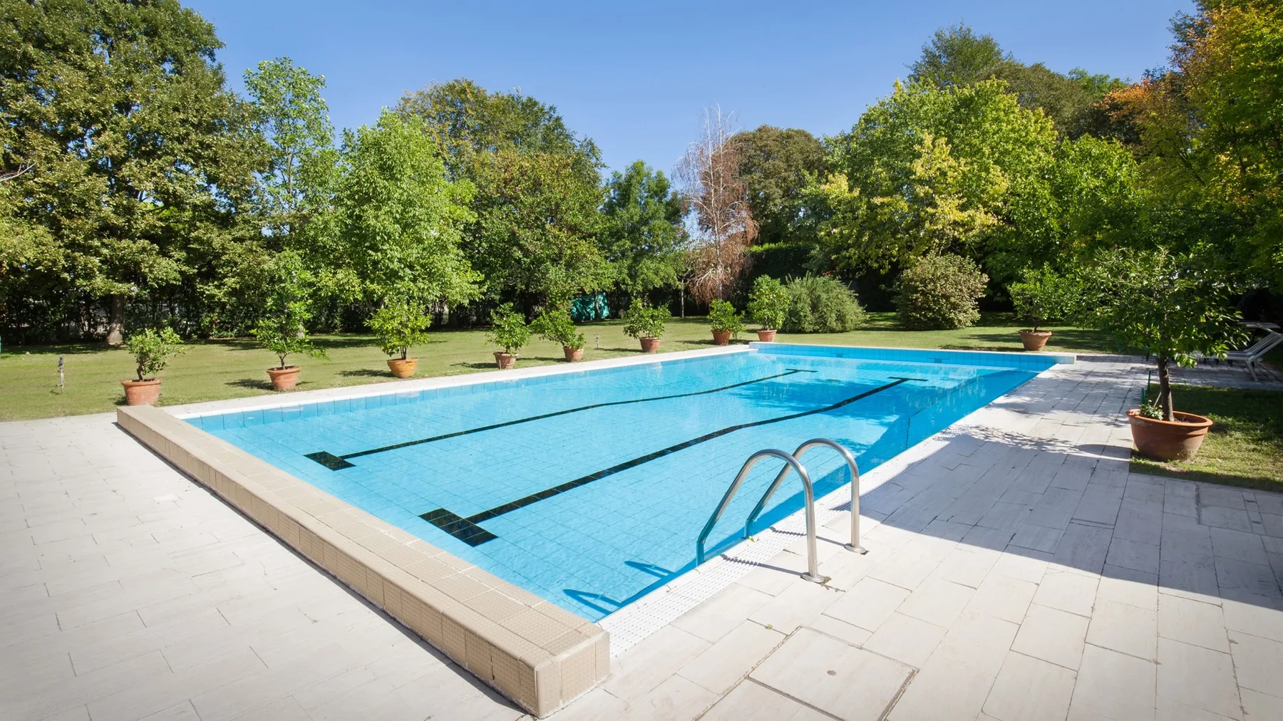 An outdoor swimming pool is surrounded by a paved deck and green trees.