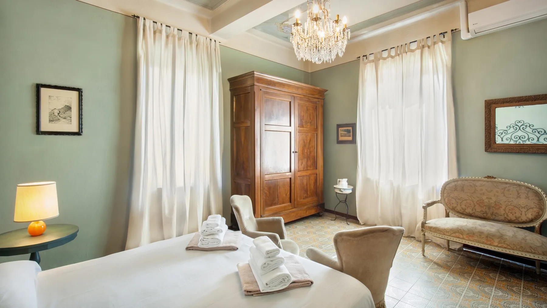 A bedroom with a wooden wardrobe, curtained windows, and a chandelier.