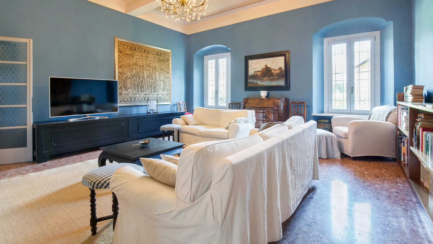 A blue living room with white seating, a television, and tall windows.