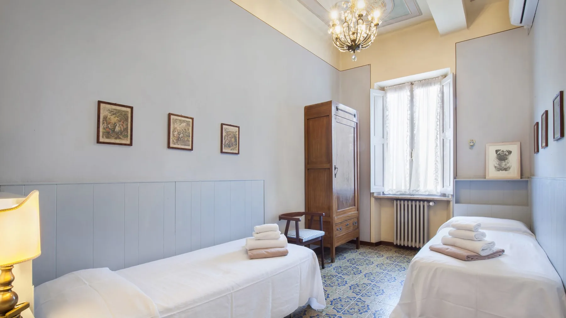 A bright bedroom with two single beds, a tall window, and patterned tile flooring.