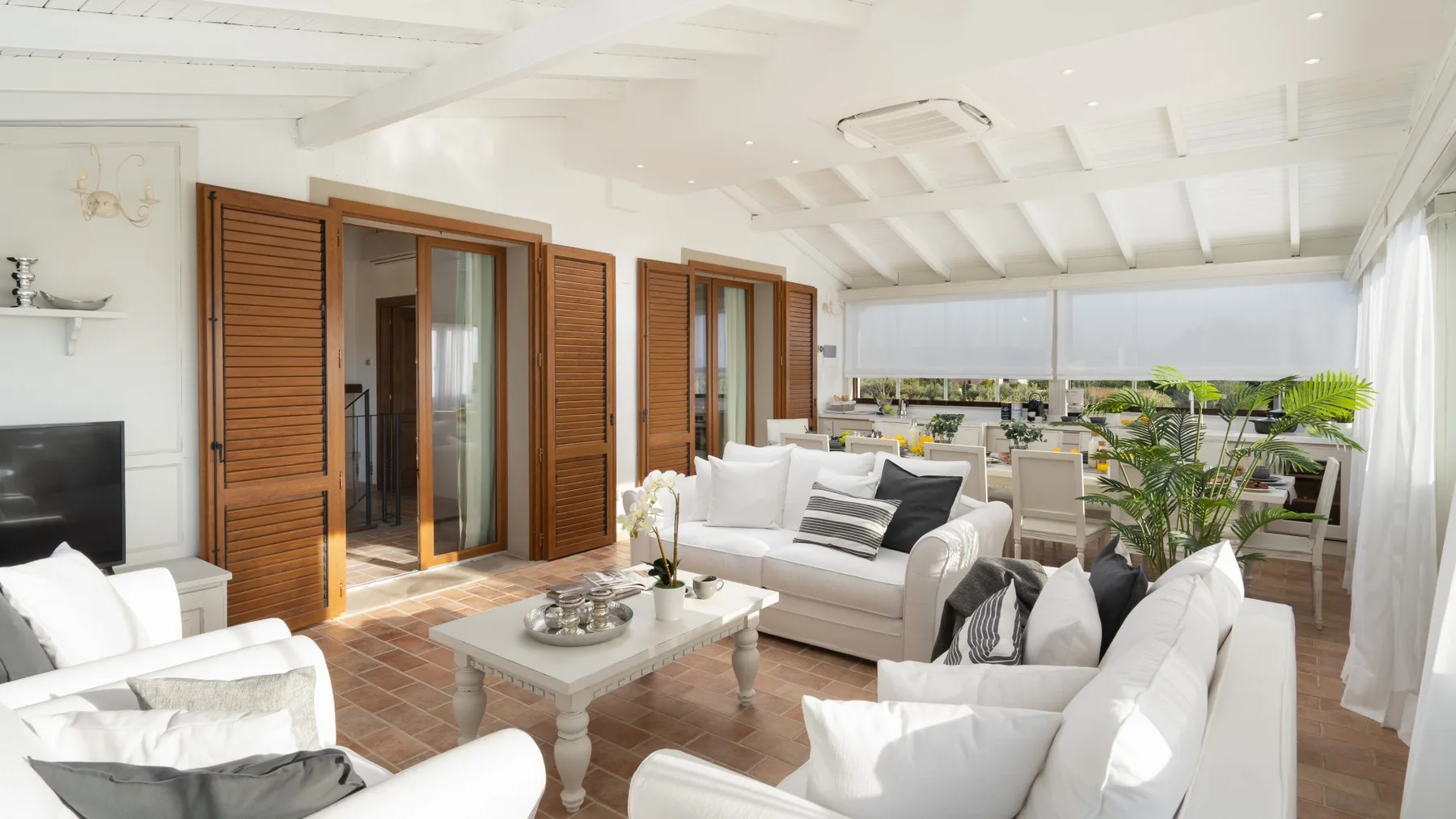 A bright living room opens onto a dining area, with white seating and large windows.