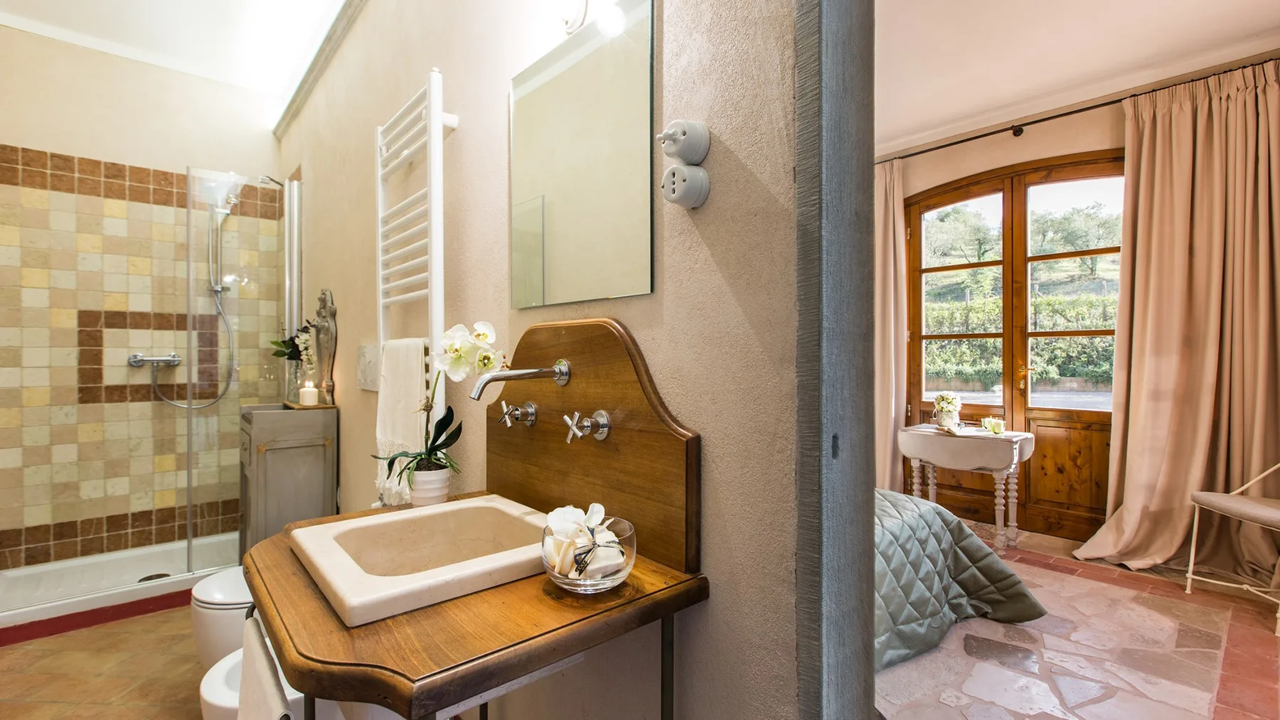 A bathroom vanity with a basin stands beside a shower and a curtained bedroom window.