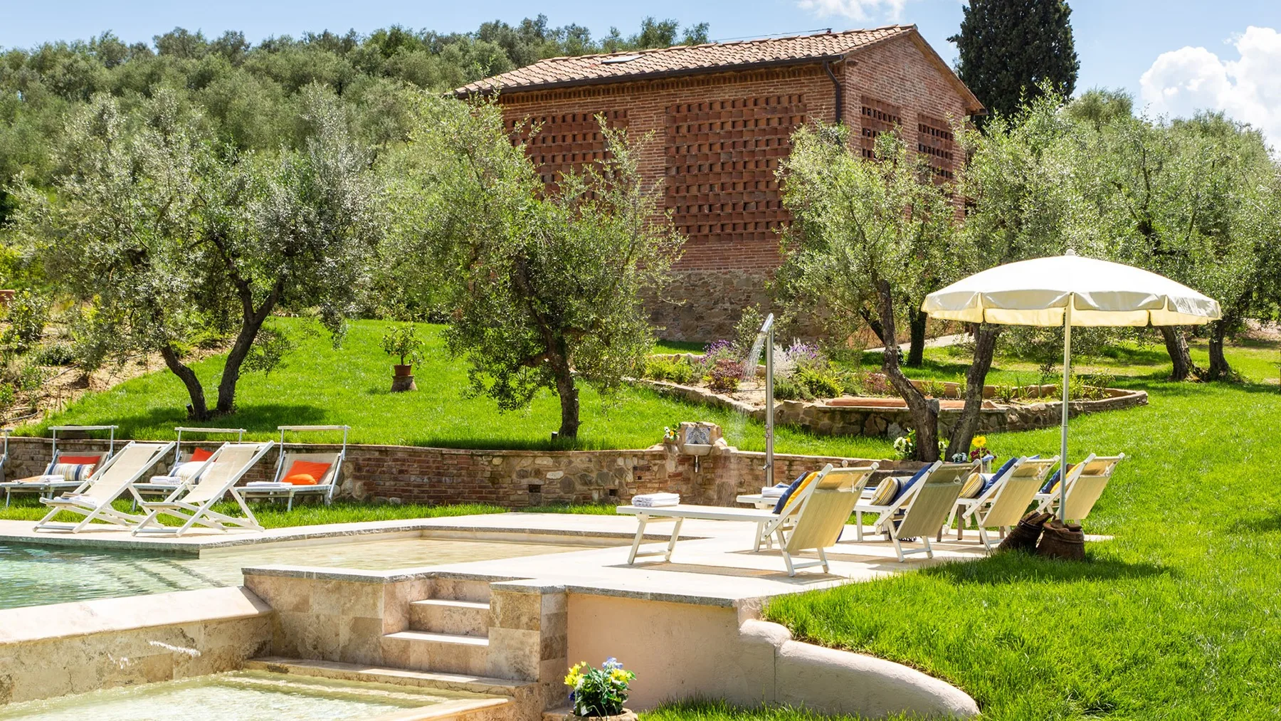An outdoor pool and sun loungers sit in a garden beneath a shade umbrella.