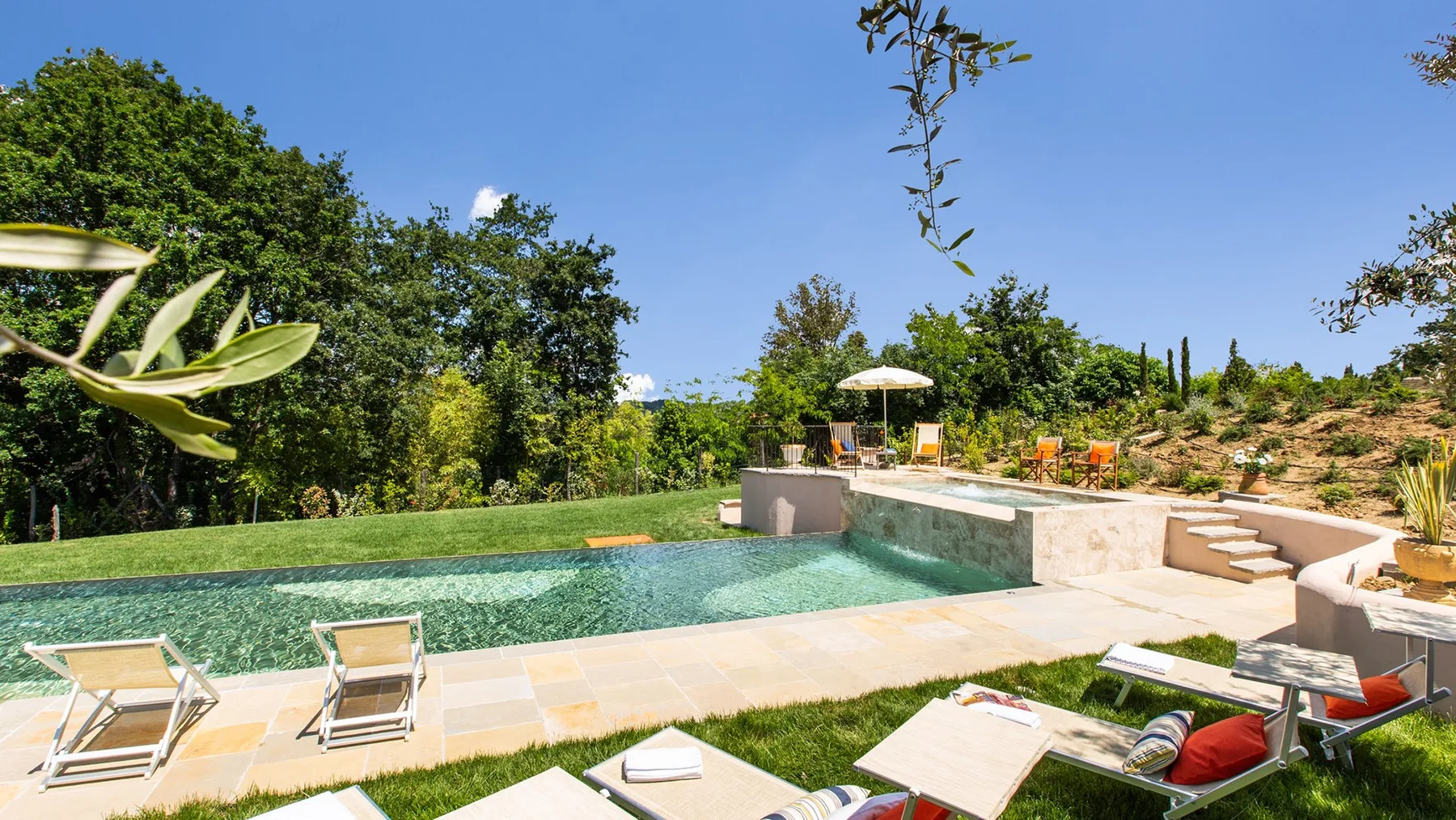 An outdoor pool beside a paved patio, lounge seating, and green landscaping.