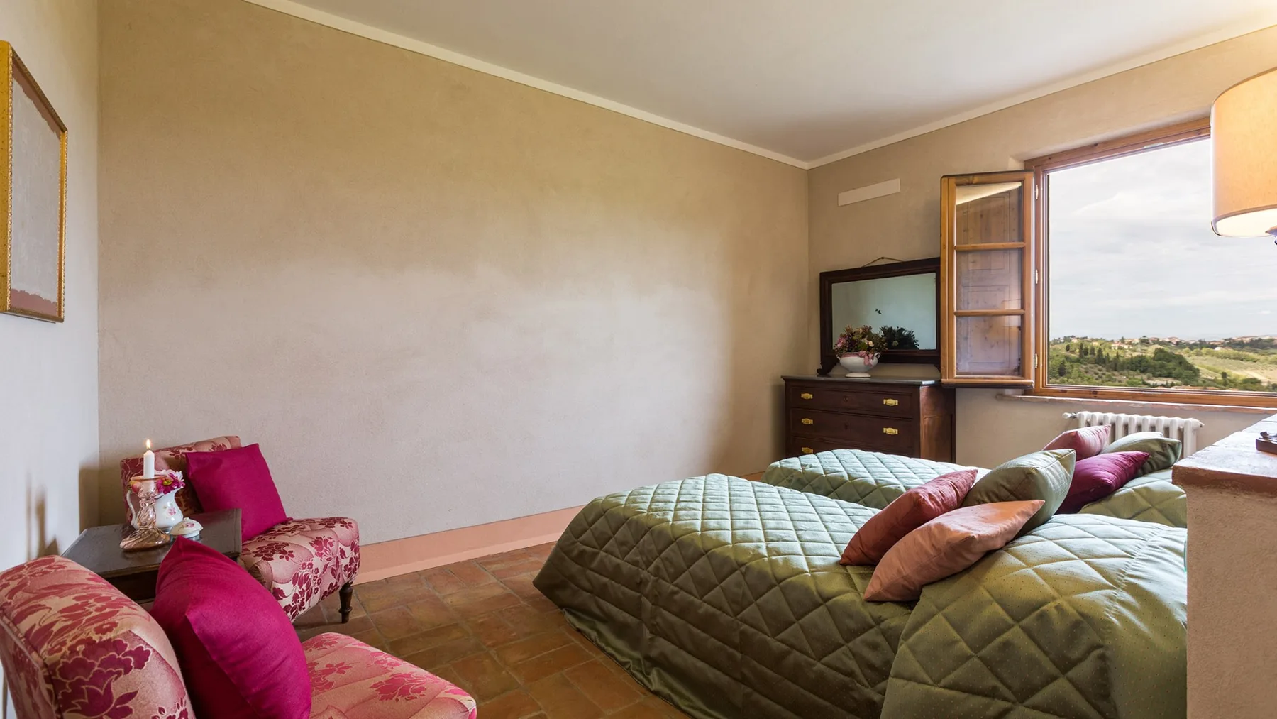 A bedroom with a large bed, patterned seating, and a window overlooking greenery.
