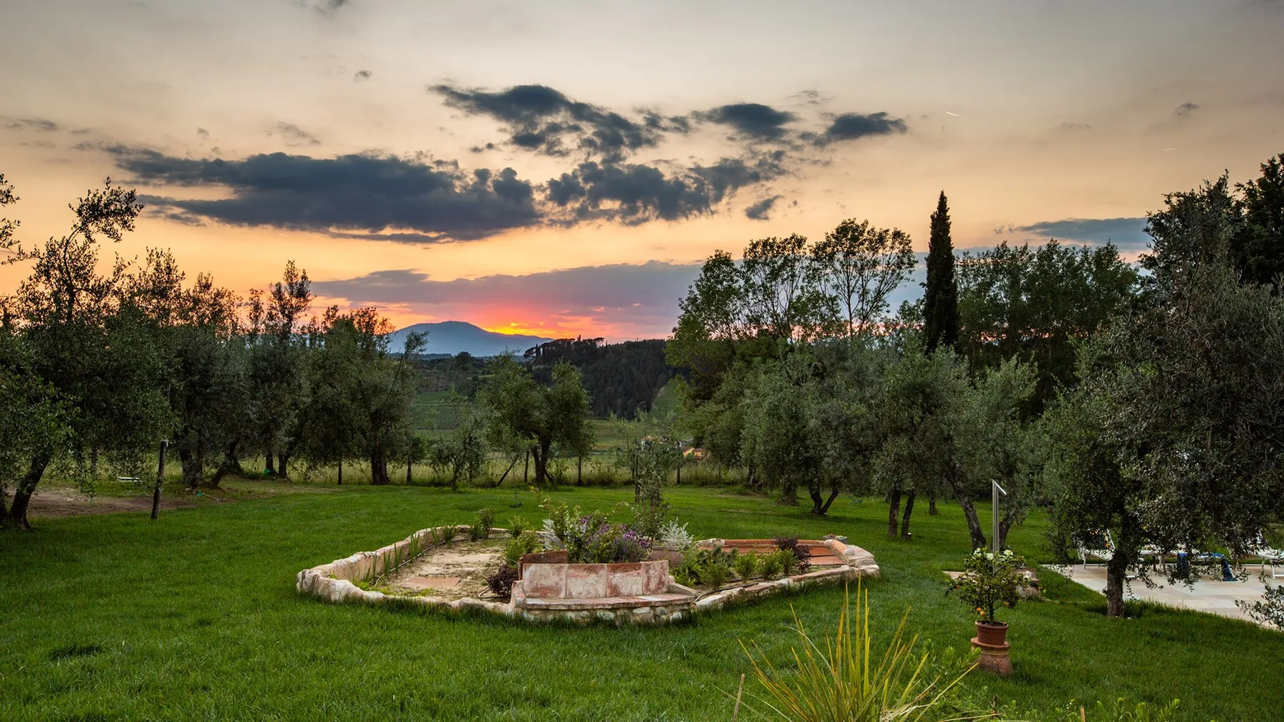 A landscaped garden with a circular stone fountain beneath a colorful sunset sky.