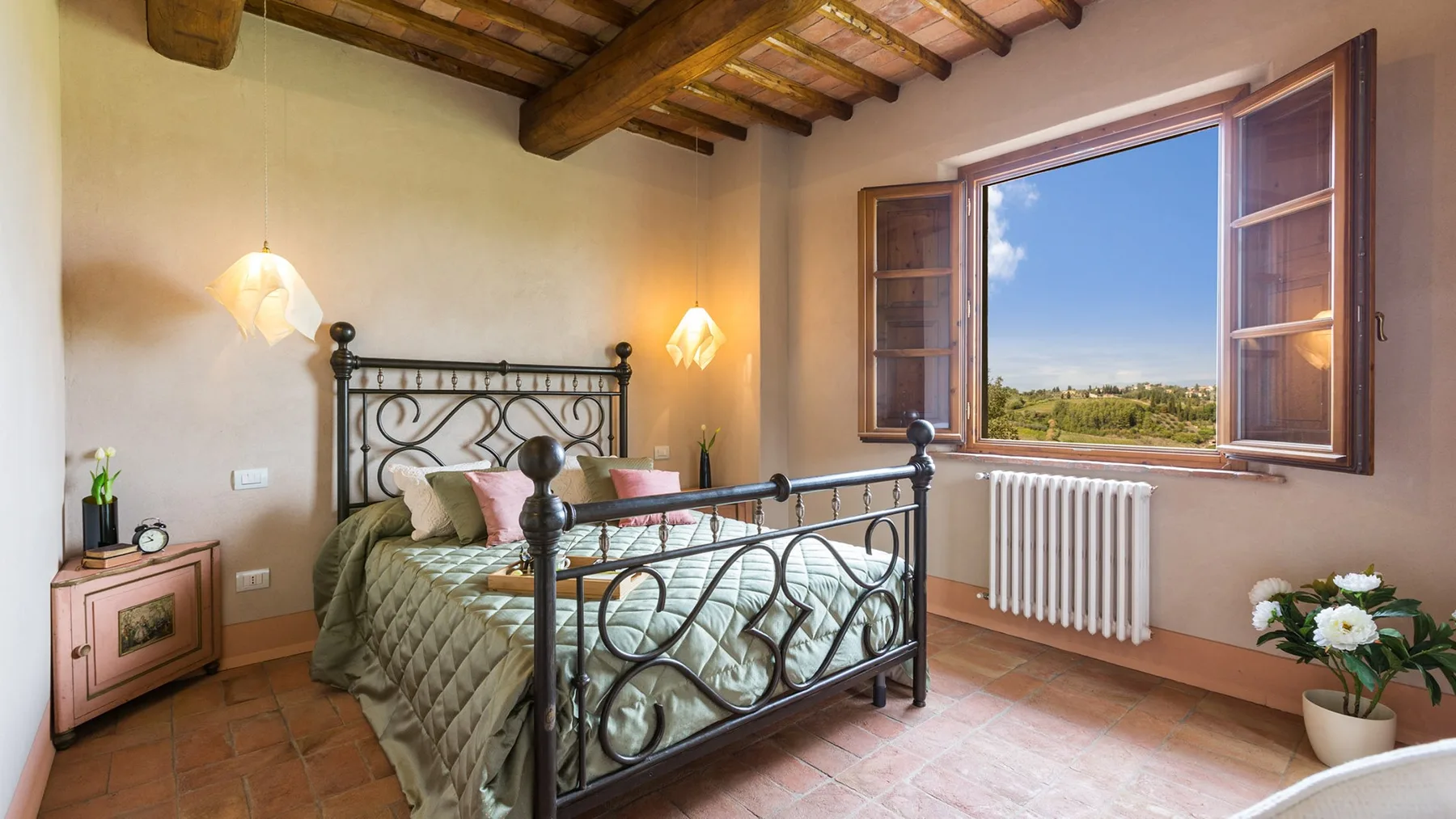 A bedroom with a metal bed, wooden ceiling beams, and an open window overlooking the countryside.
