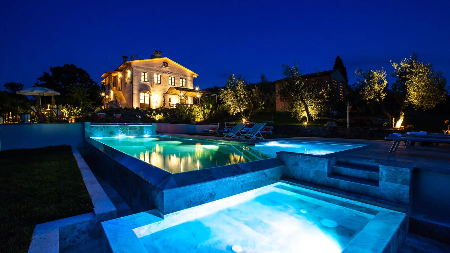 An illuminated outdoor swimming pool sits in front of a warmly lit building at night.