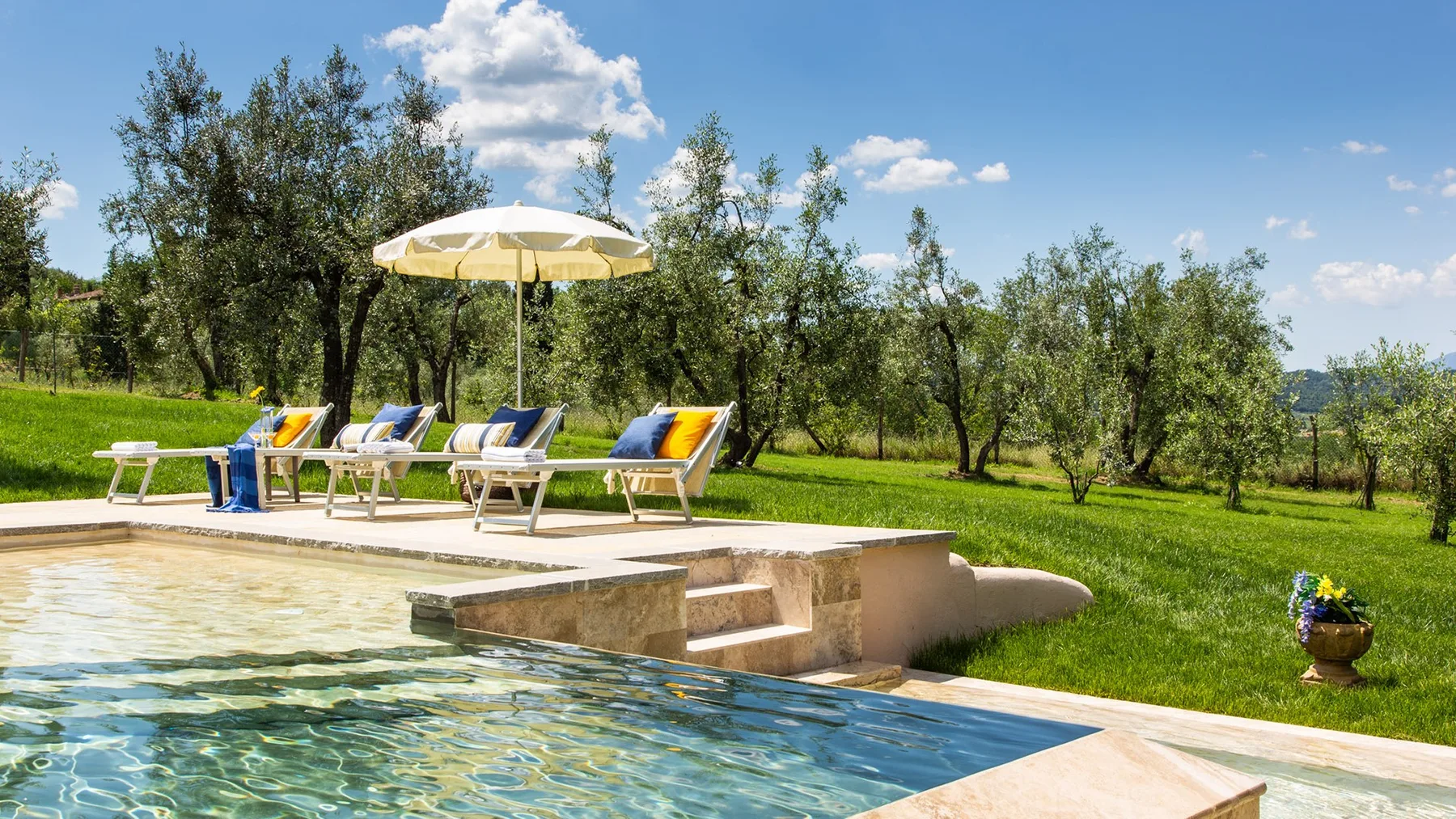 An outdoor swimming pool beside a sunny patio with lounge seating and an umbrella.