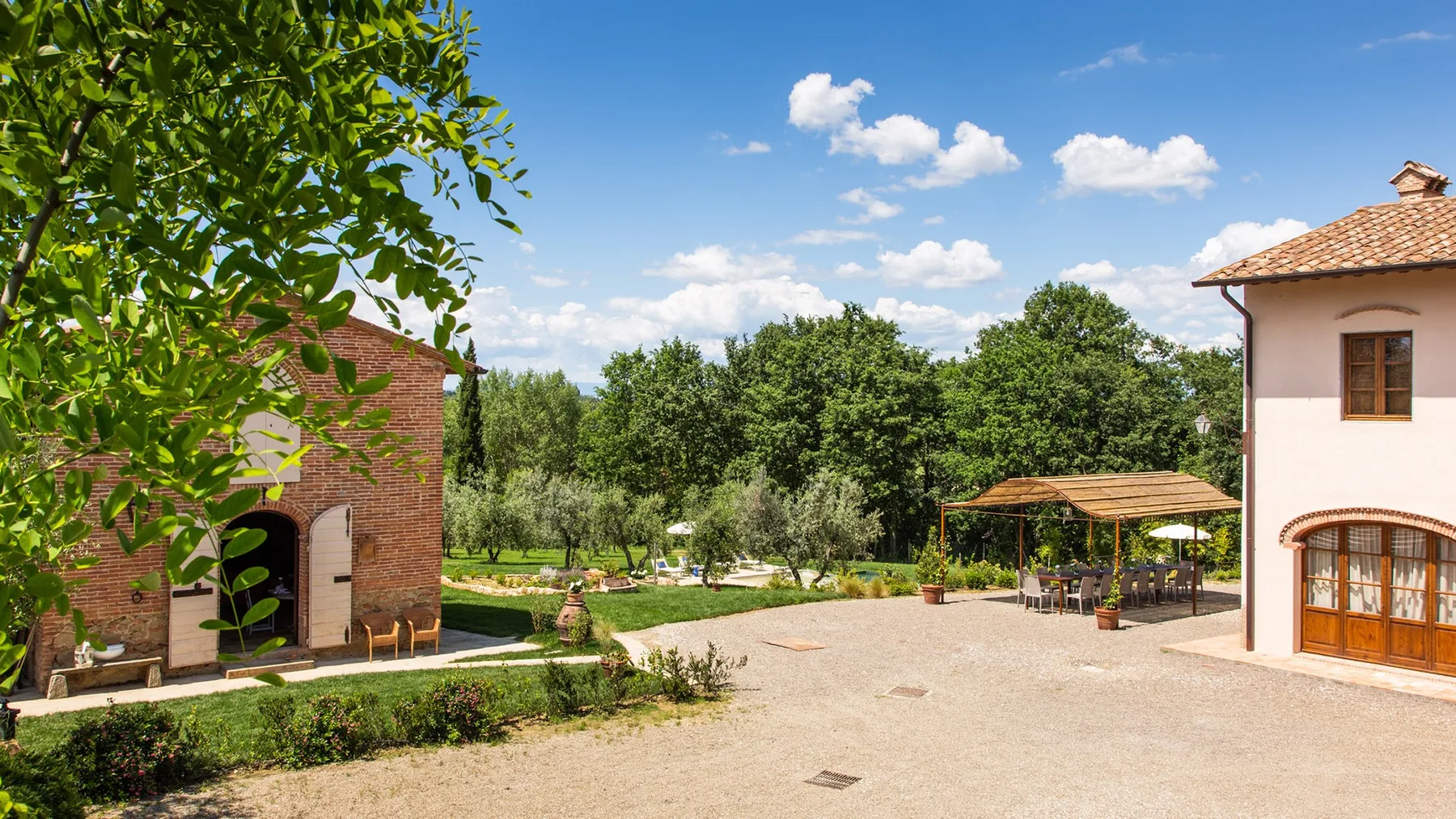 A sunny outdoor courtyard sits between two buildings, with trees and a covered seating area.