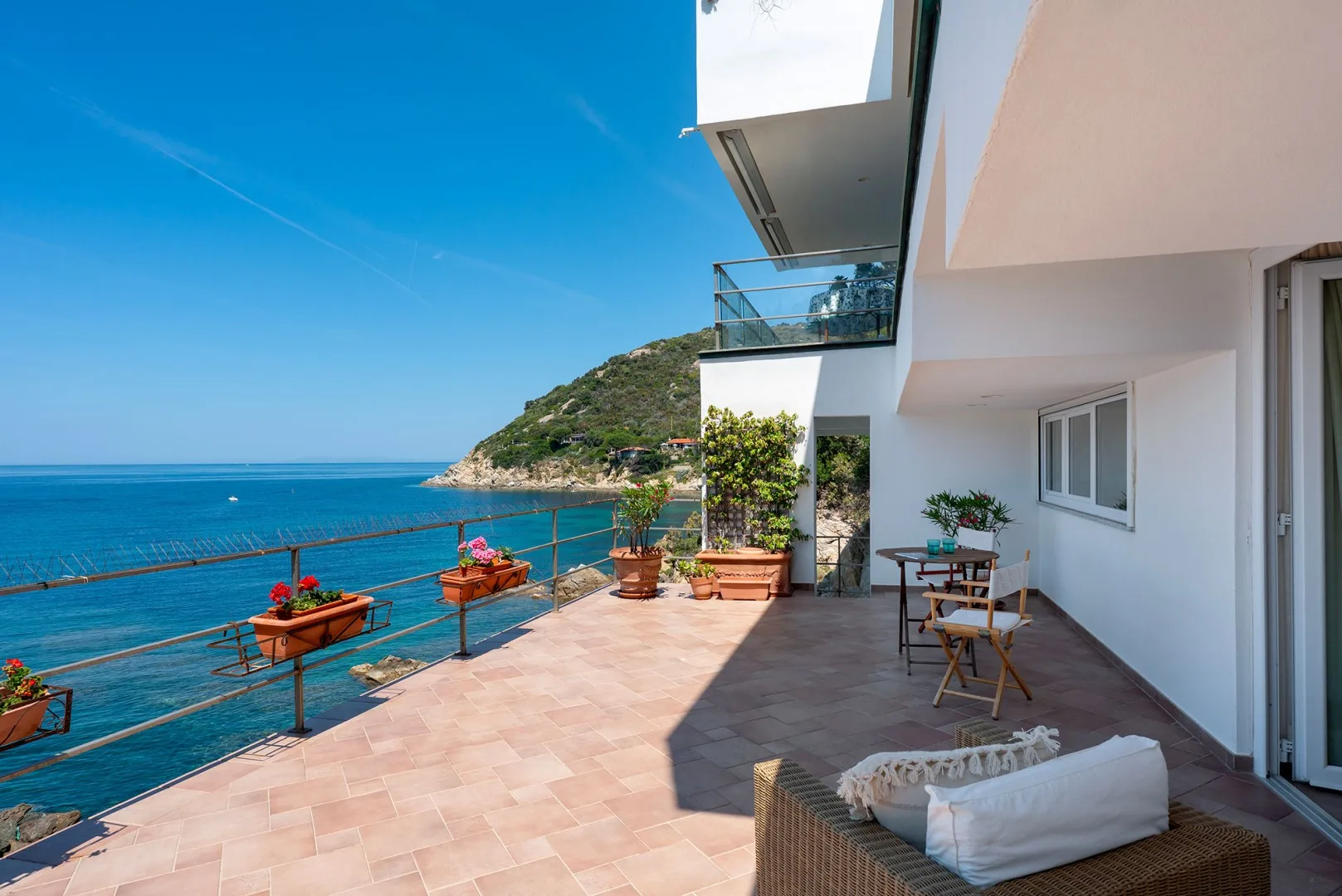 A sunny seaside terrace with outdoor seating overlooking the blue water.