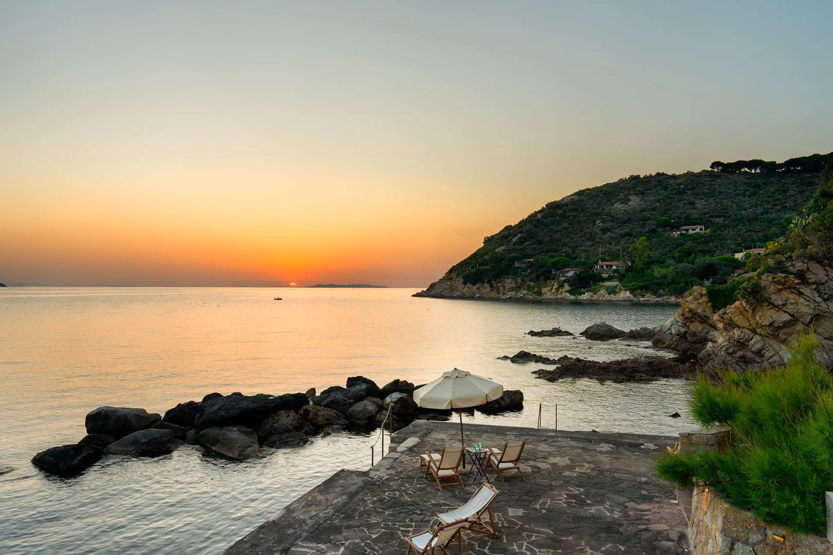 A rocky seaside terrace overlooks calm water and a sunset.