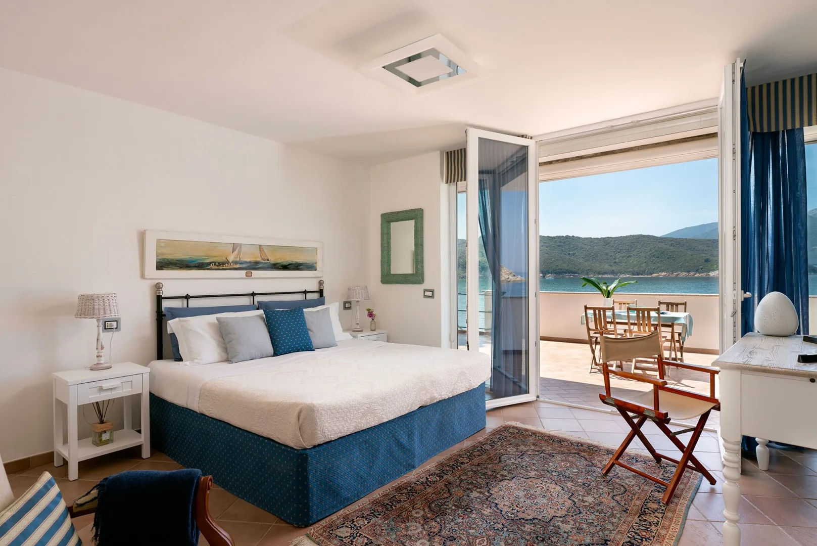 A bedroom with a bed beside open doors leading to a balcony overlooking the water.