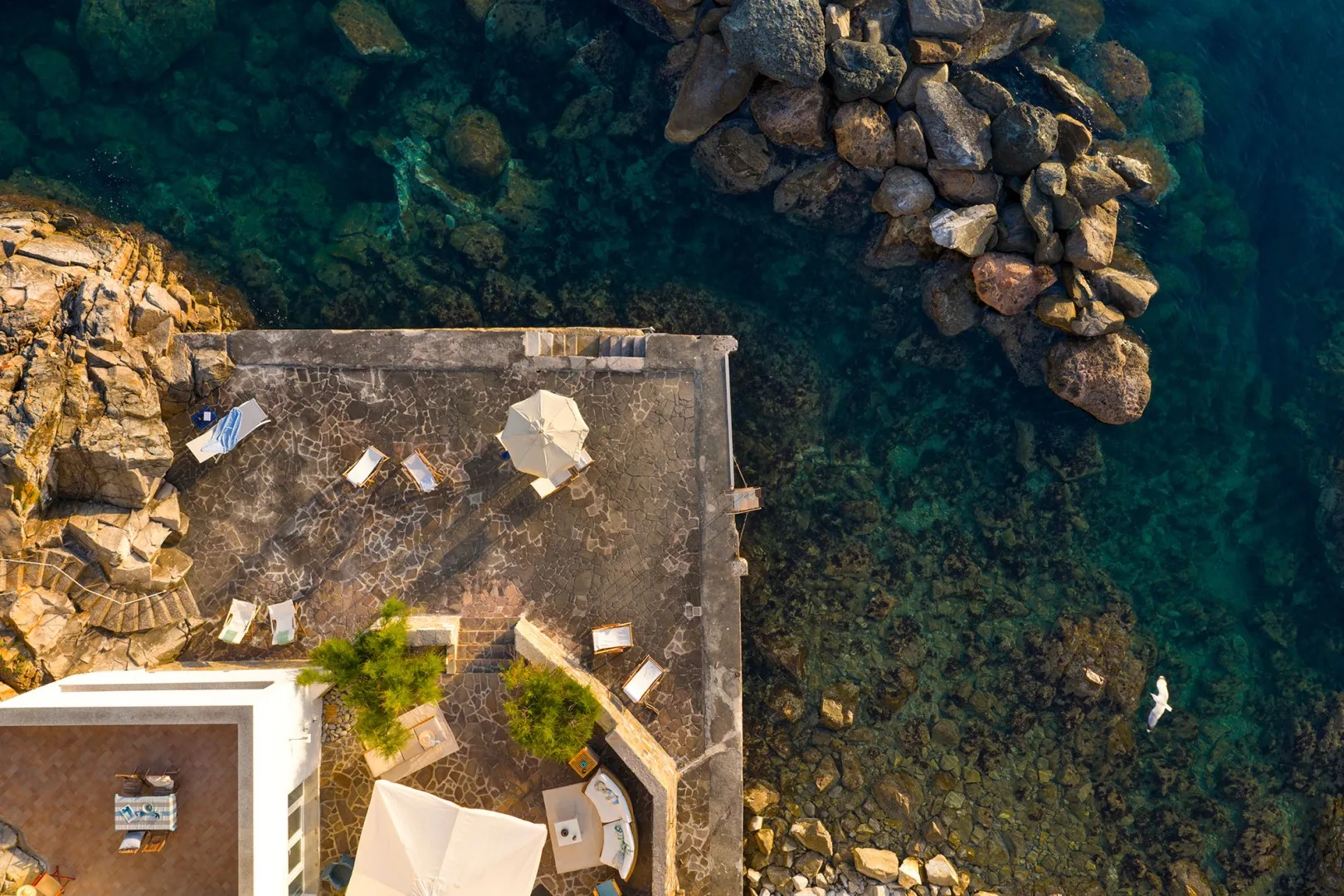 An overhead view shows a seaside terrace beside clear water and rocky shoreline.
