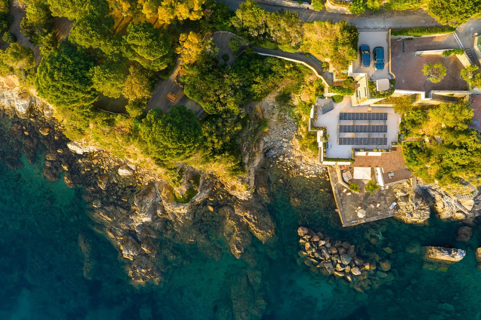 An aerial view shows a coastal property beside rocky shoreline and turquoise water.