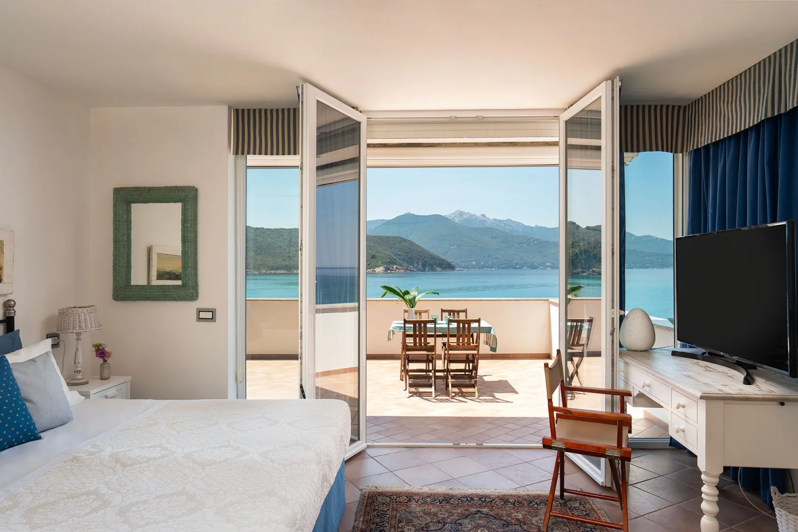 A bedroom opens onto a terrace with a dining table and a view of the water and mountains.
