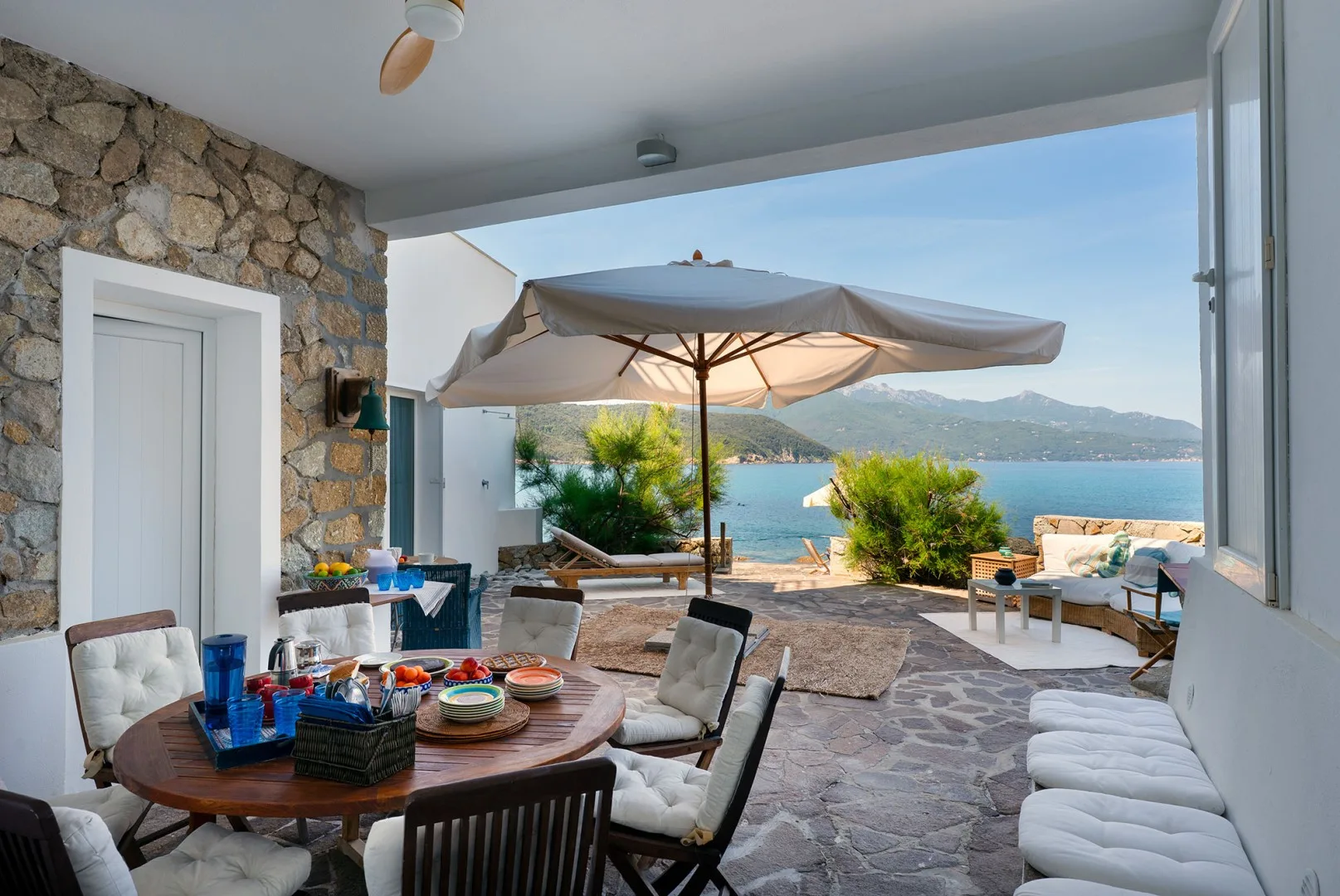 A shaded seaside patio has outdoor seating and a large umbrella overlooking the water.