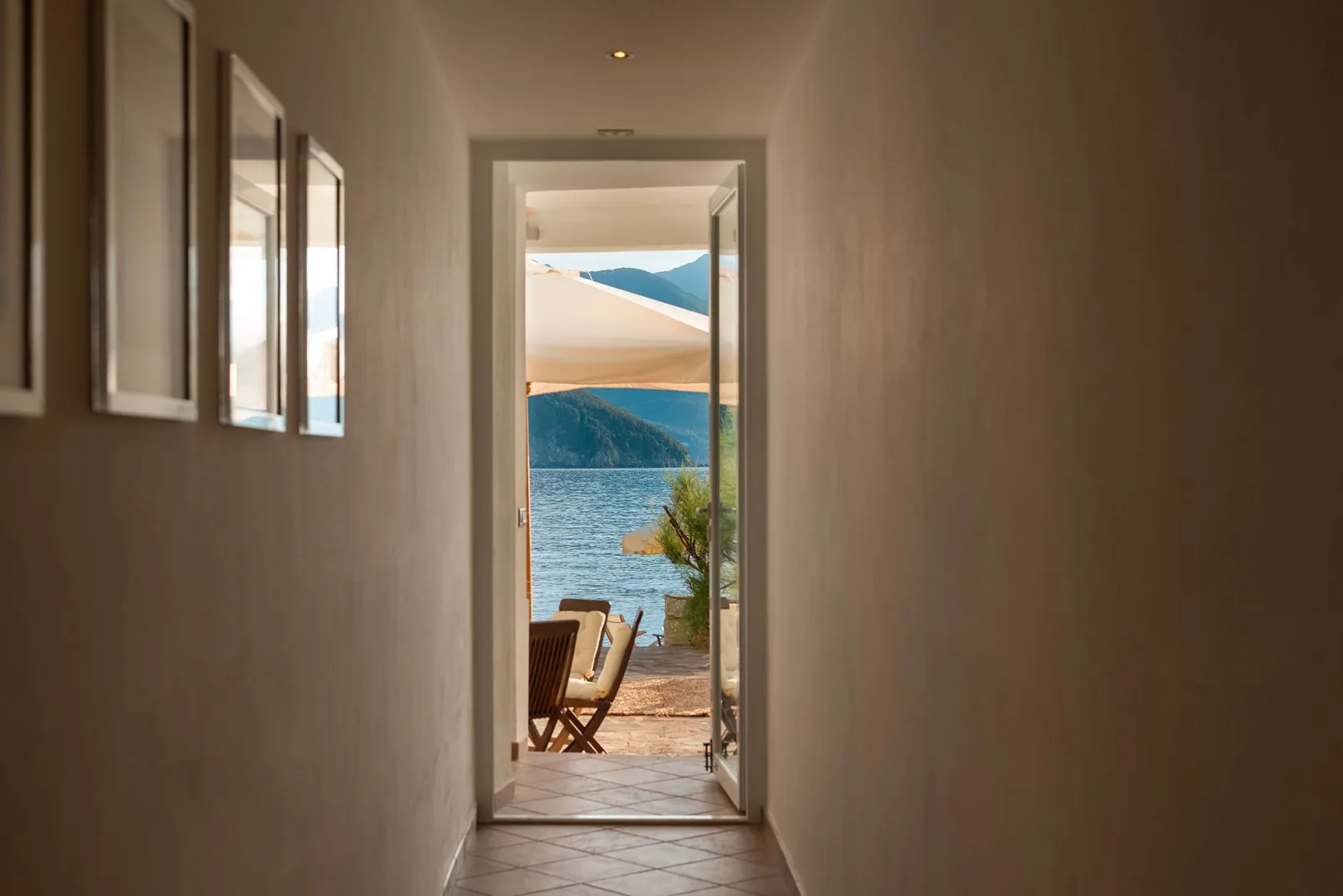 A hallway opens onto a waterside terrace with seating and a mountain view.