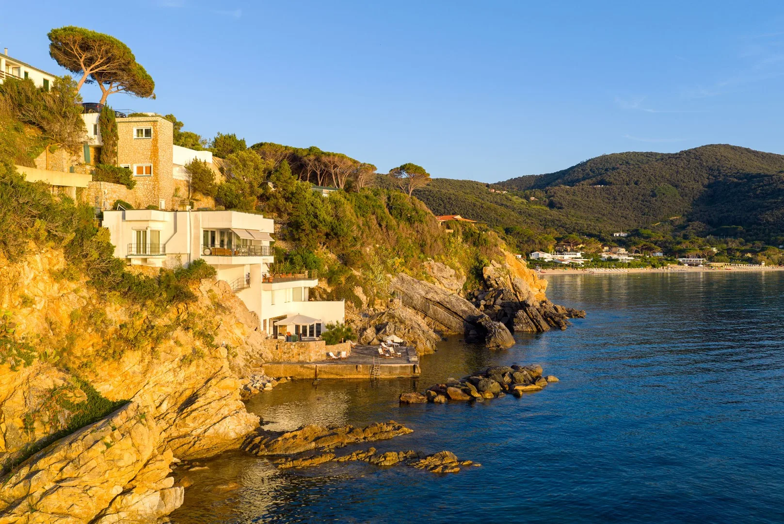 Coastal homes sit above rocky cliffs beside calm blue water.