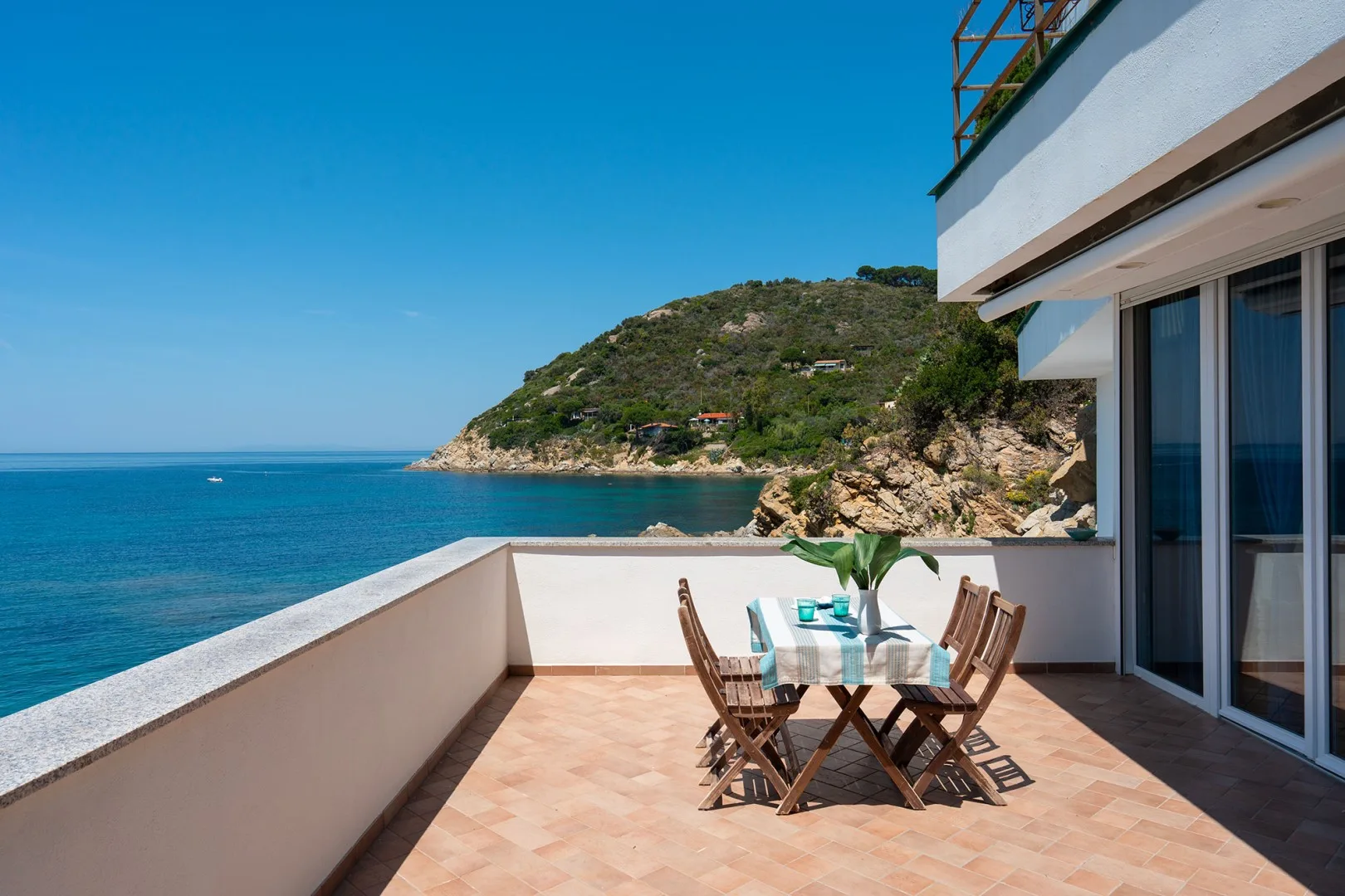 A seaside terrace with outdoor seating overlooks the blue water and a green headland.
