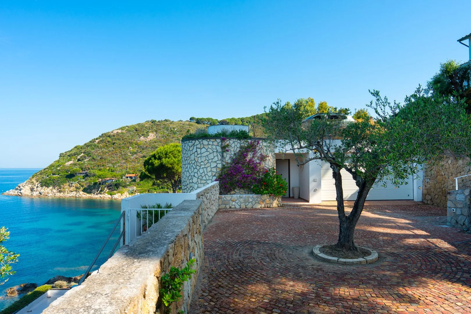 A seaside courtyard with stone paving, a tree, and a view of the blue water.