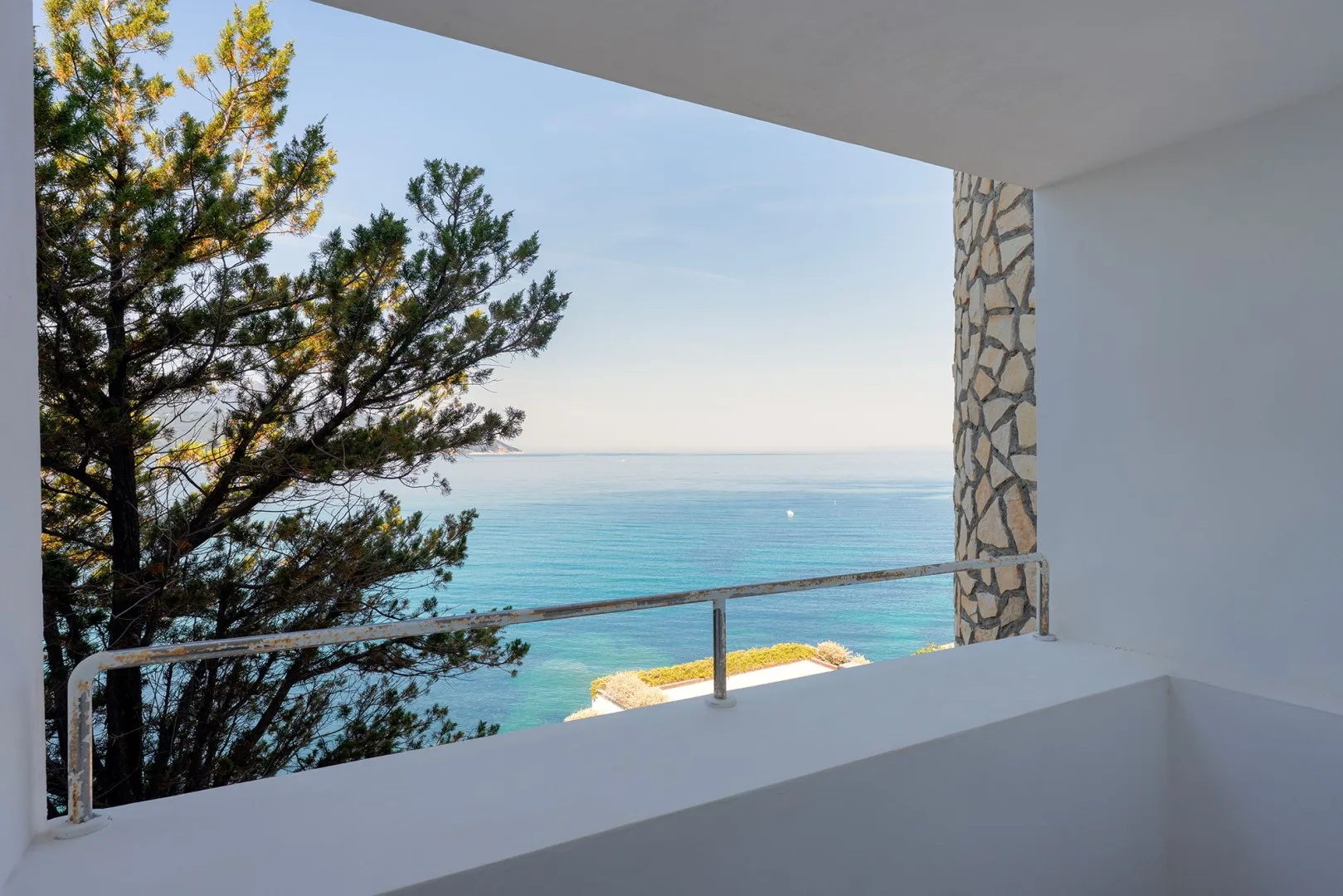 A covered balcony overlooks the turquoise sea, with a tree beside the railing.