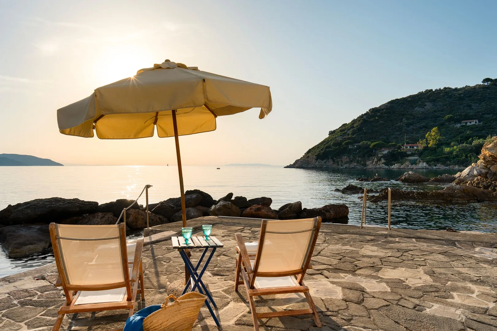 A seaside patio with two lounge chairs and an umbrella overlooking calm water.