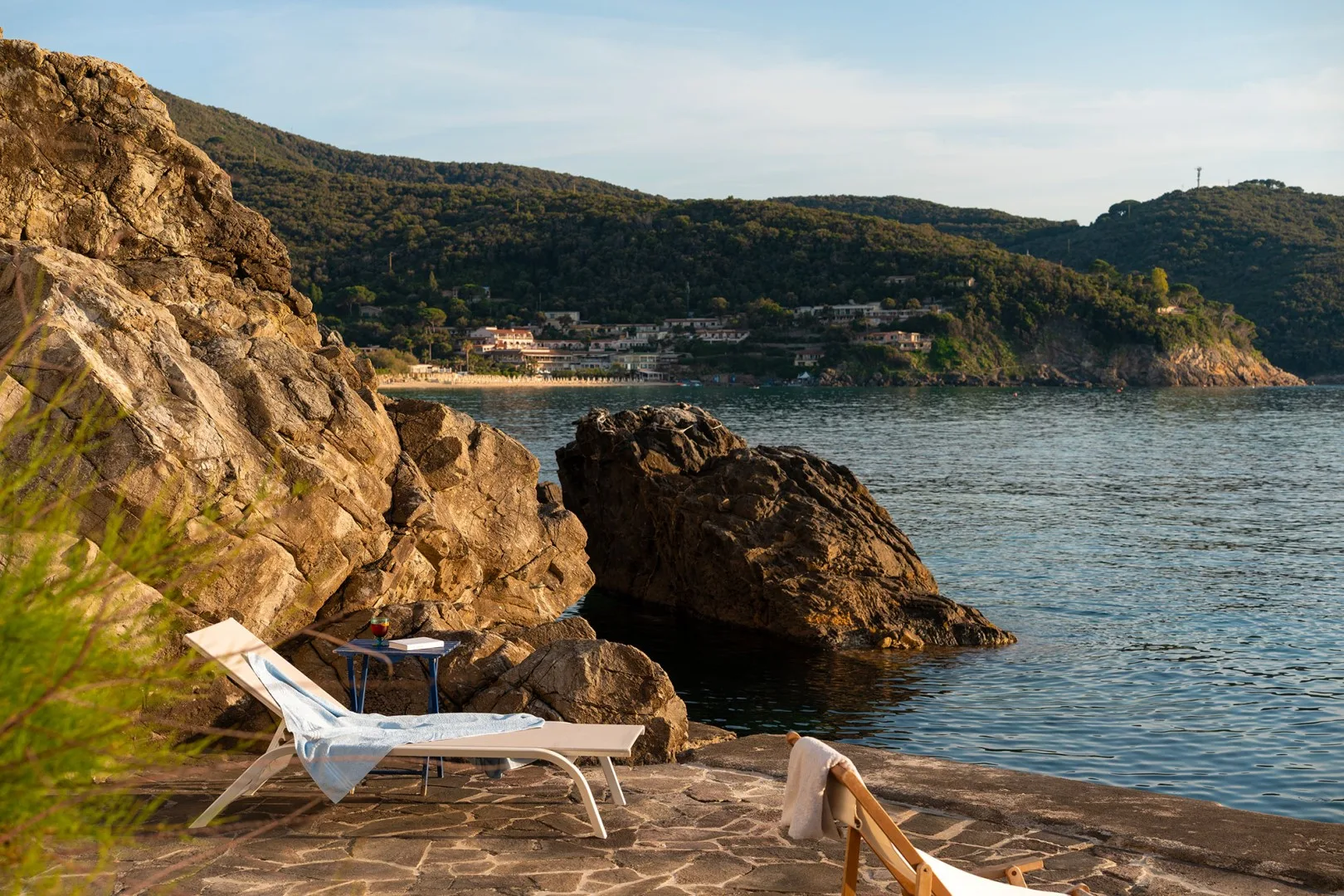 A rocky seaside terrace overlooks the water, with a lounge chair near the shore.