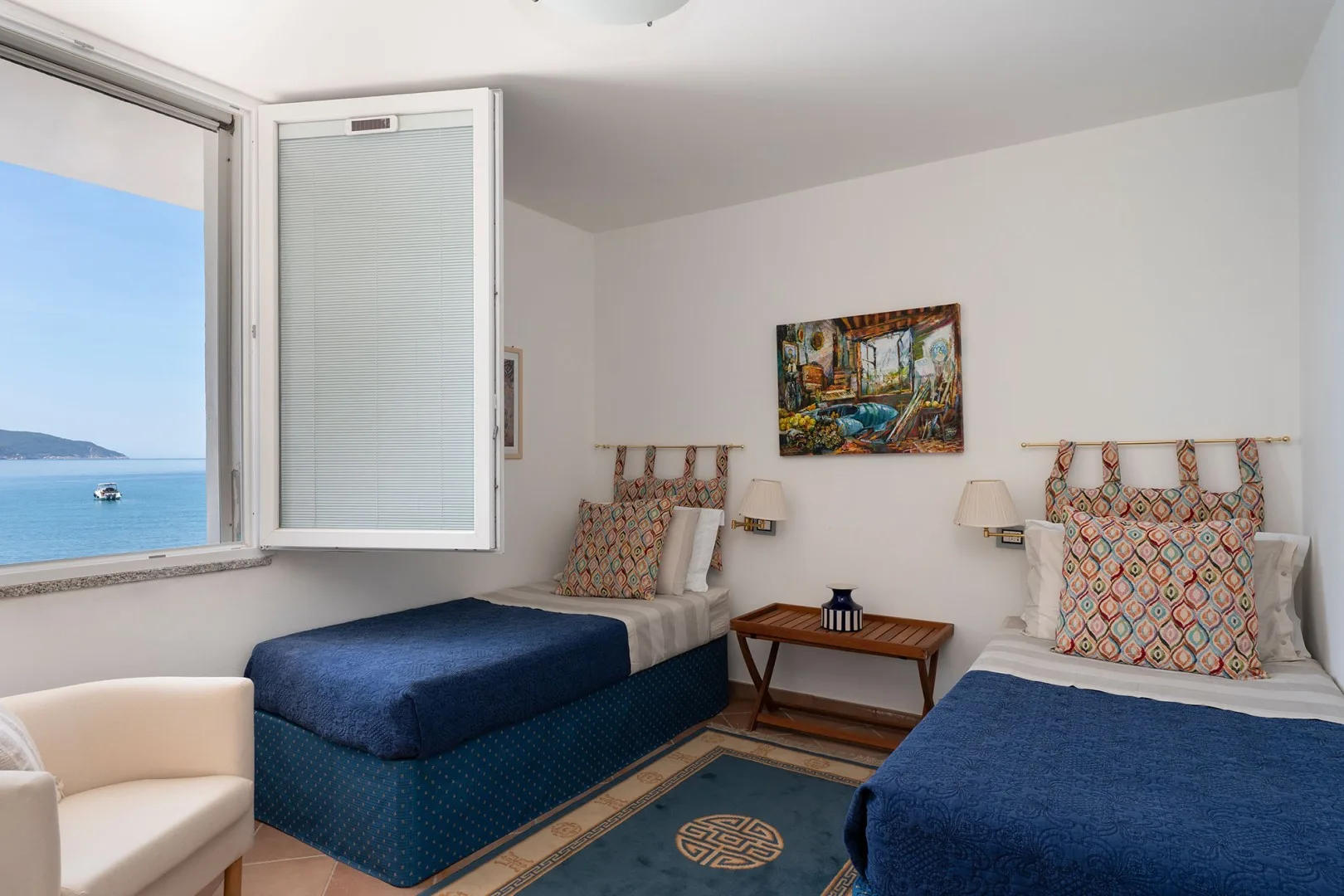 A bedroom with two beds, patterned pillows, and an open window overlooking the water.