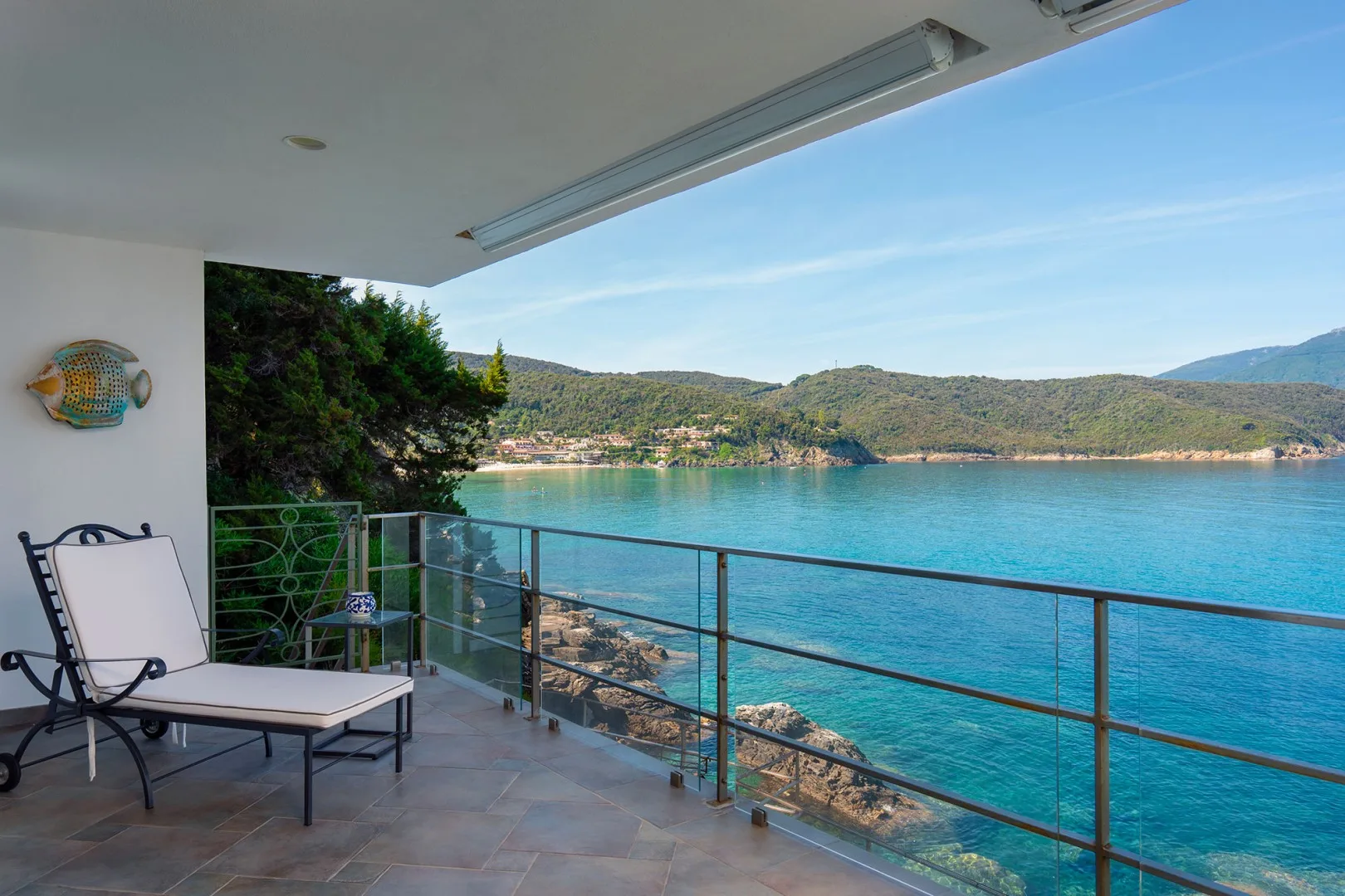 A balcony with a lounge chair overlooks a turquoise bay and wooded hills.