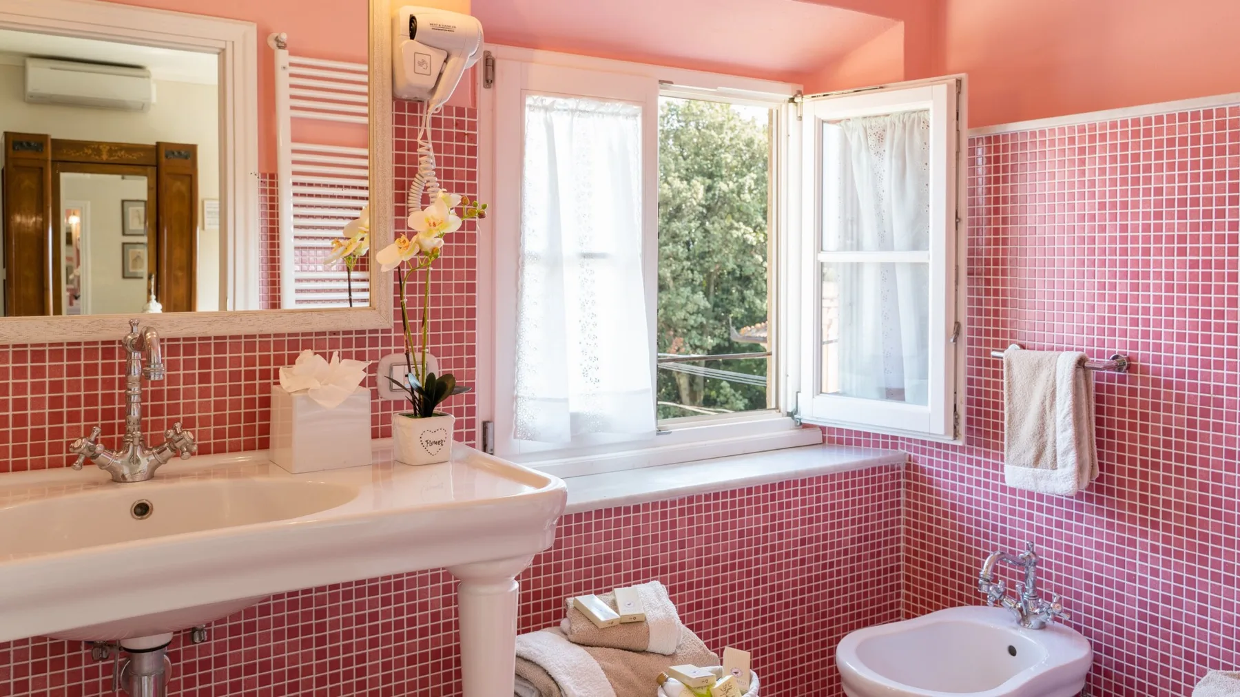 Un baño con azulejos rosas, lavabo, bidé y una ventana abierta.